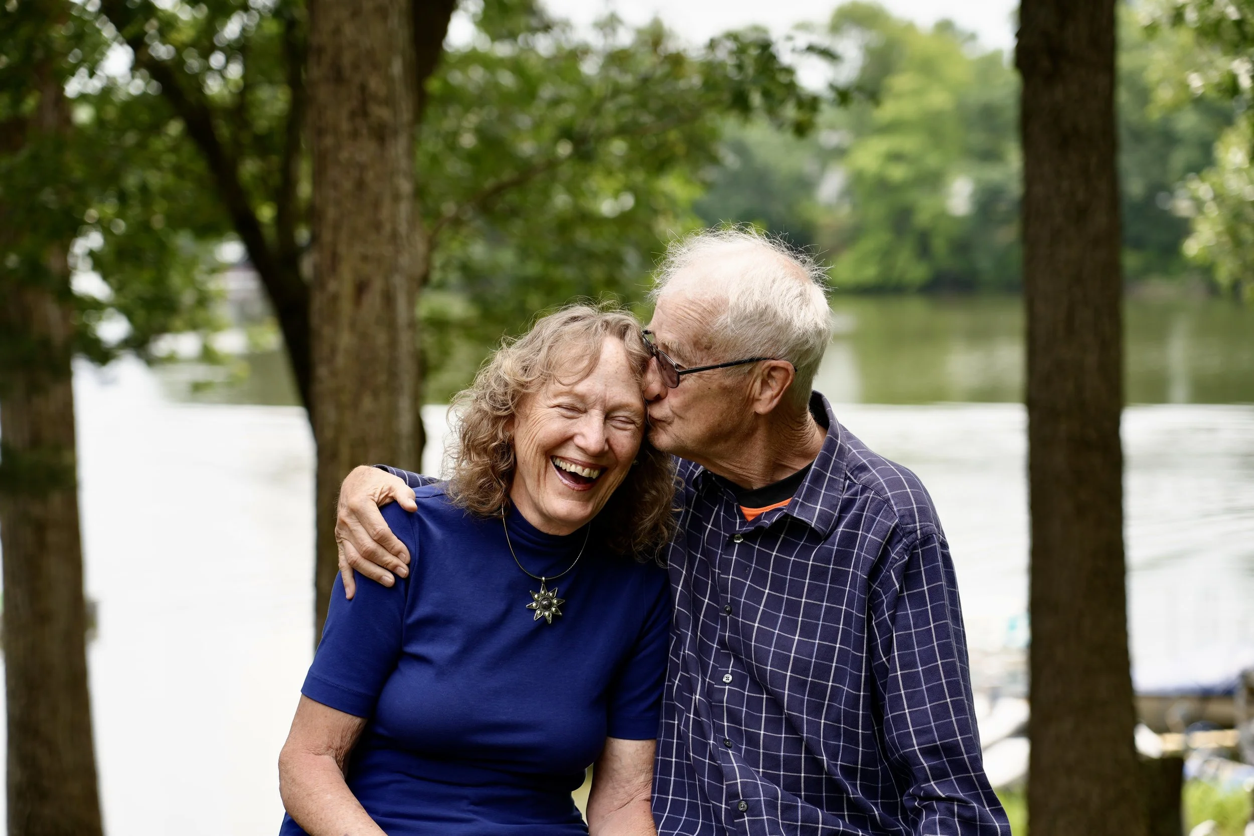 An older couple sharing a joyful moment in a park, with the man kissing the woman's cheek. They are seated near a lake, surrounded by trees.