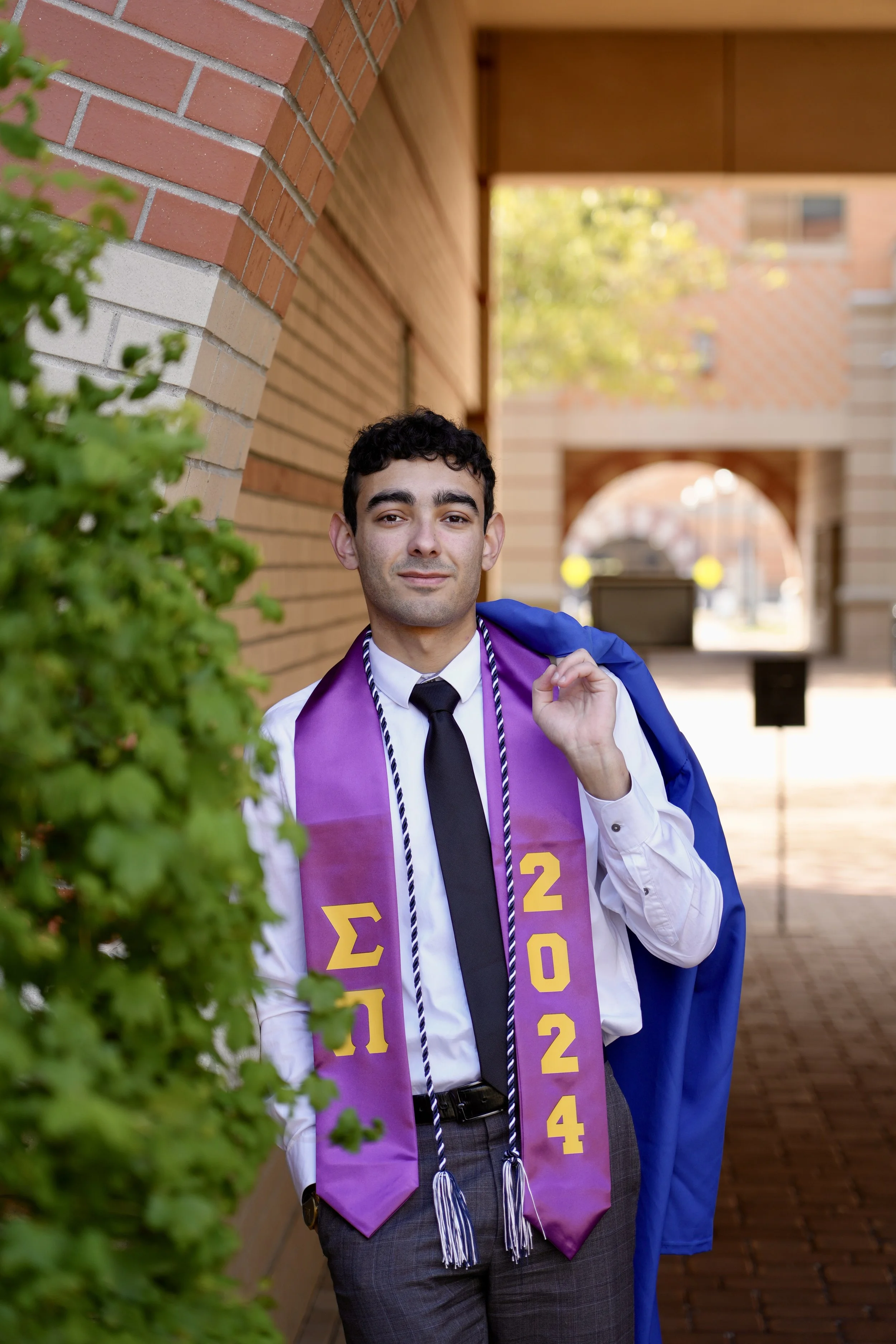 Graduate wearing a purple stole with Greek letters and "2024", holding a blue graduation gown, standing by a brick wall, with greenery in the foreground.