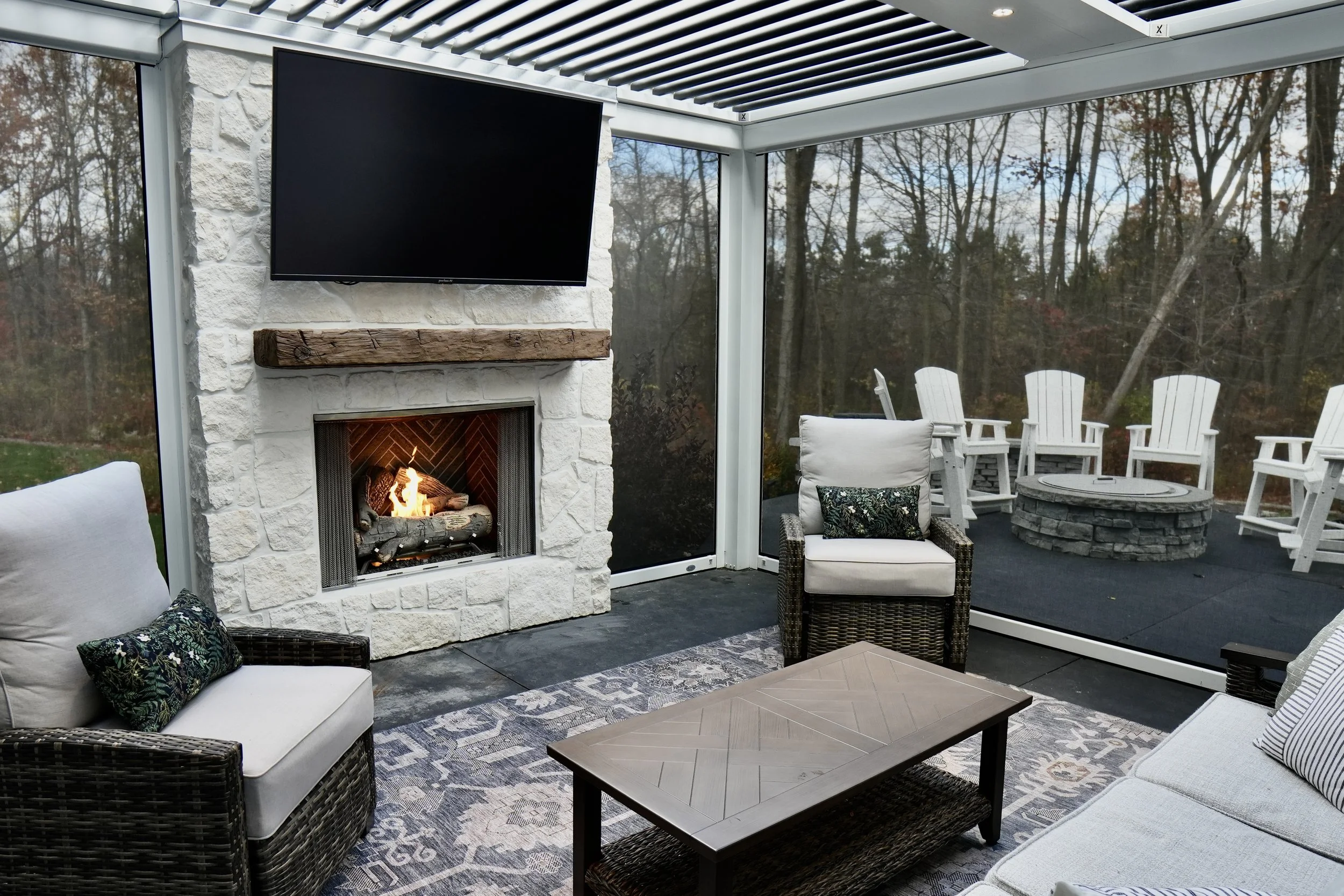 Cozy outdoor patio with wicker chairs, a fireplace, a mounted TV, and surrounding trees.