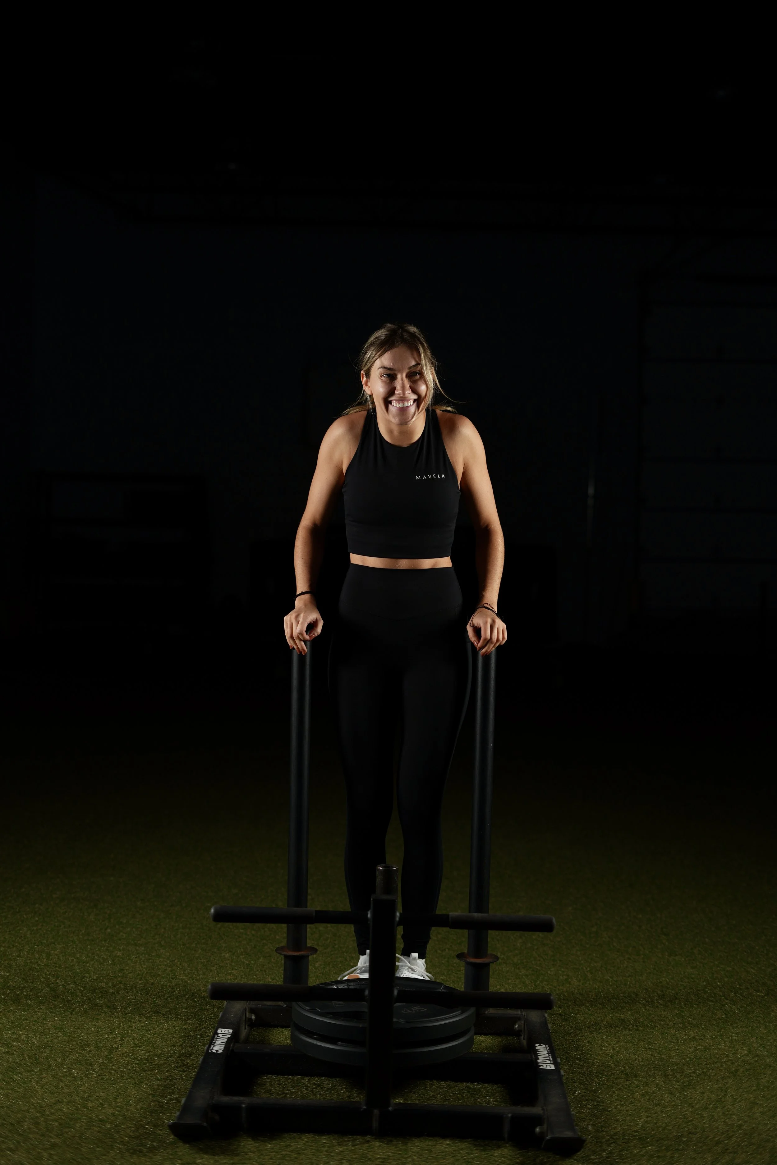 A woman in black athletic wear smiling and standing on a sled push machine in a dark indoor gym.