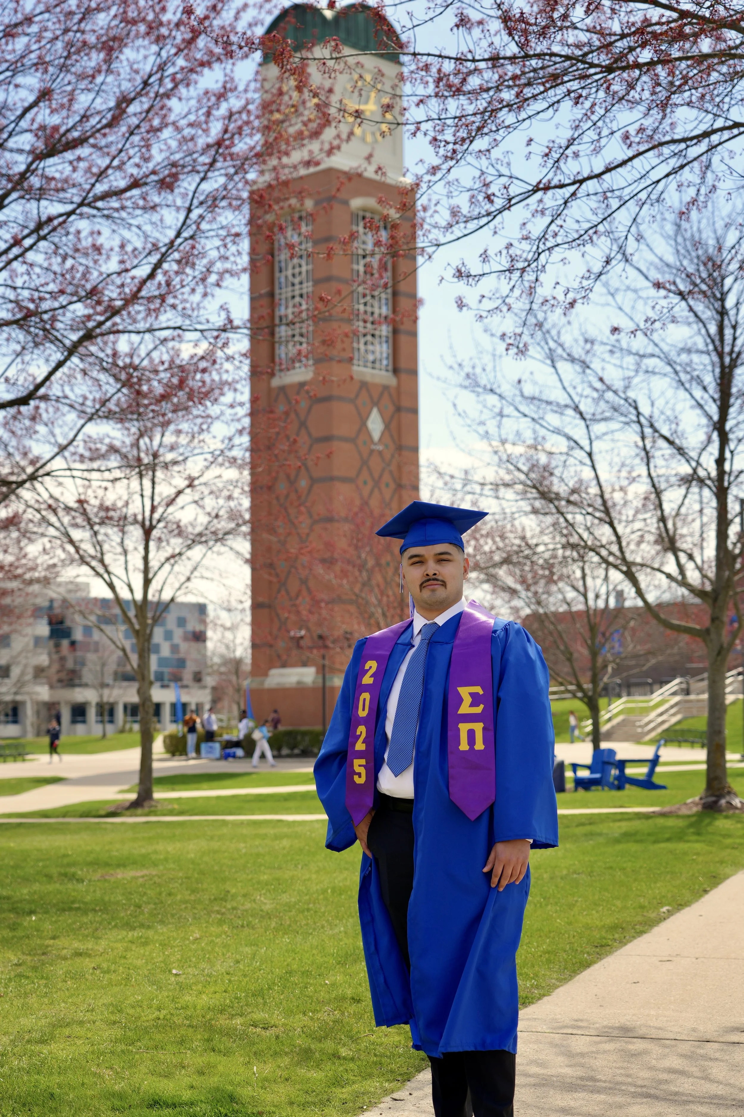 A man in a blue graduation gown and cap standing outdoors on a university campus with a clock tower in the background, pink flowering trees, and other students in the distance.