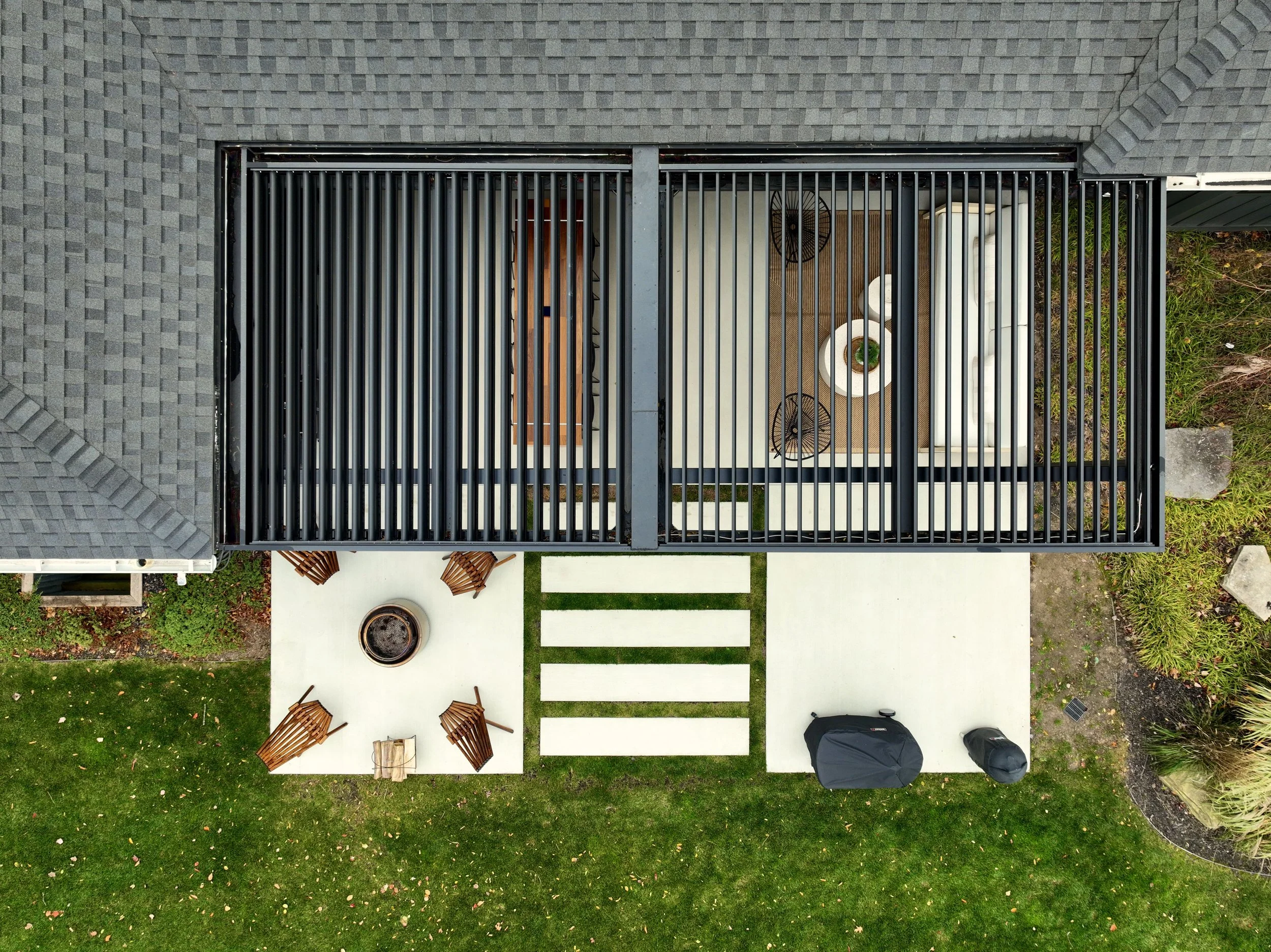 Aerial view of a modern backyard with a black rooftop pergola, outdoor furniture, a fire pit surrounded by chairs, a grill, and a lawn with trees.