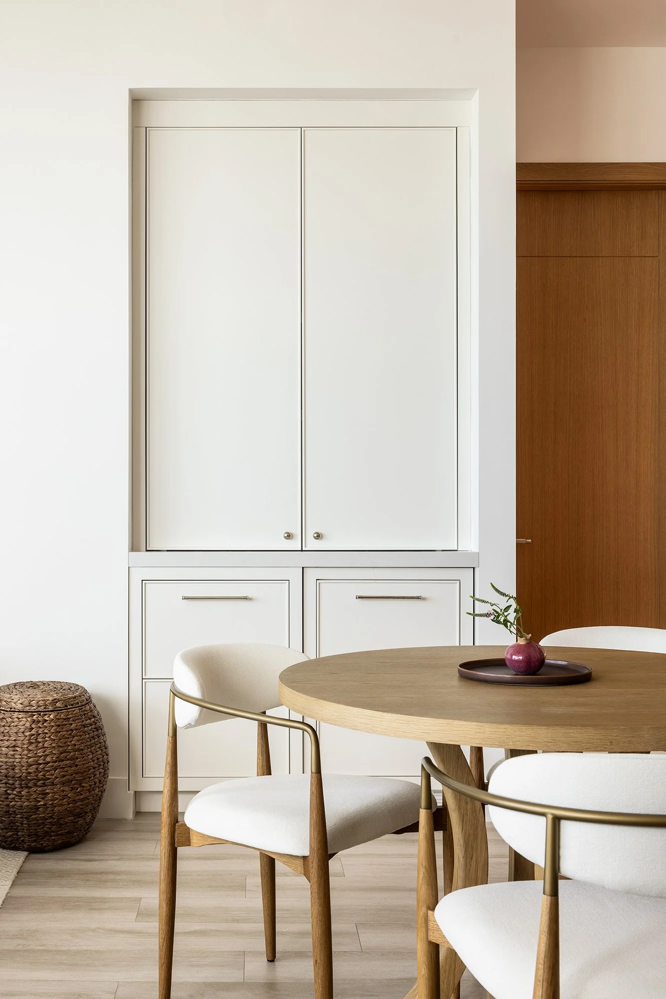 Modern dining area with a round wooden table, white chairs with wooden legs, and a small decorative item on the table. A white cabinet with closed doors is mounted on the wall behind the table, next to a wooden partition.