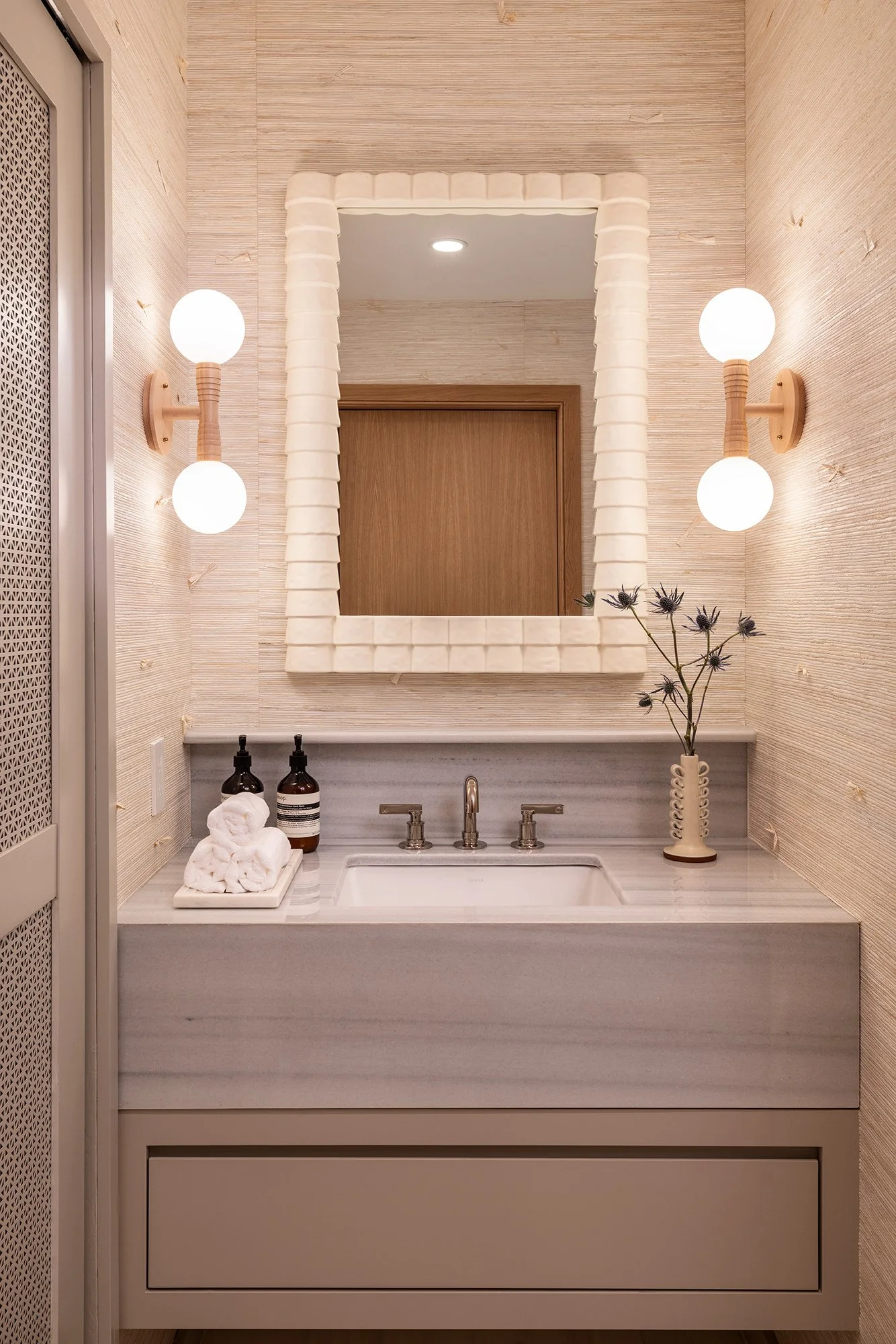 A bathroom vanity with a white marble countertop, a rectangular sink, and modern fixtures. The mirror has a textured white frame, flanked by two wall-mounted light fixtures with frosted globes and wooden accents. To the right of the sink is a decorative vase with dried flowers, and on the left are bottles of soap and a bundle of rolled white towels.