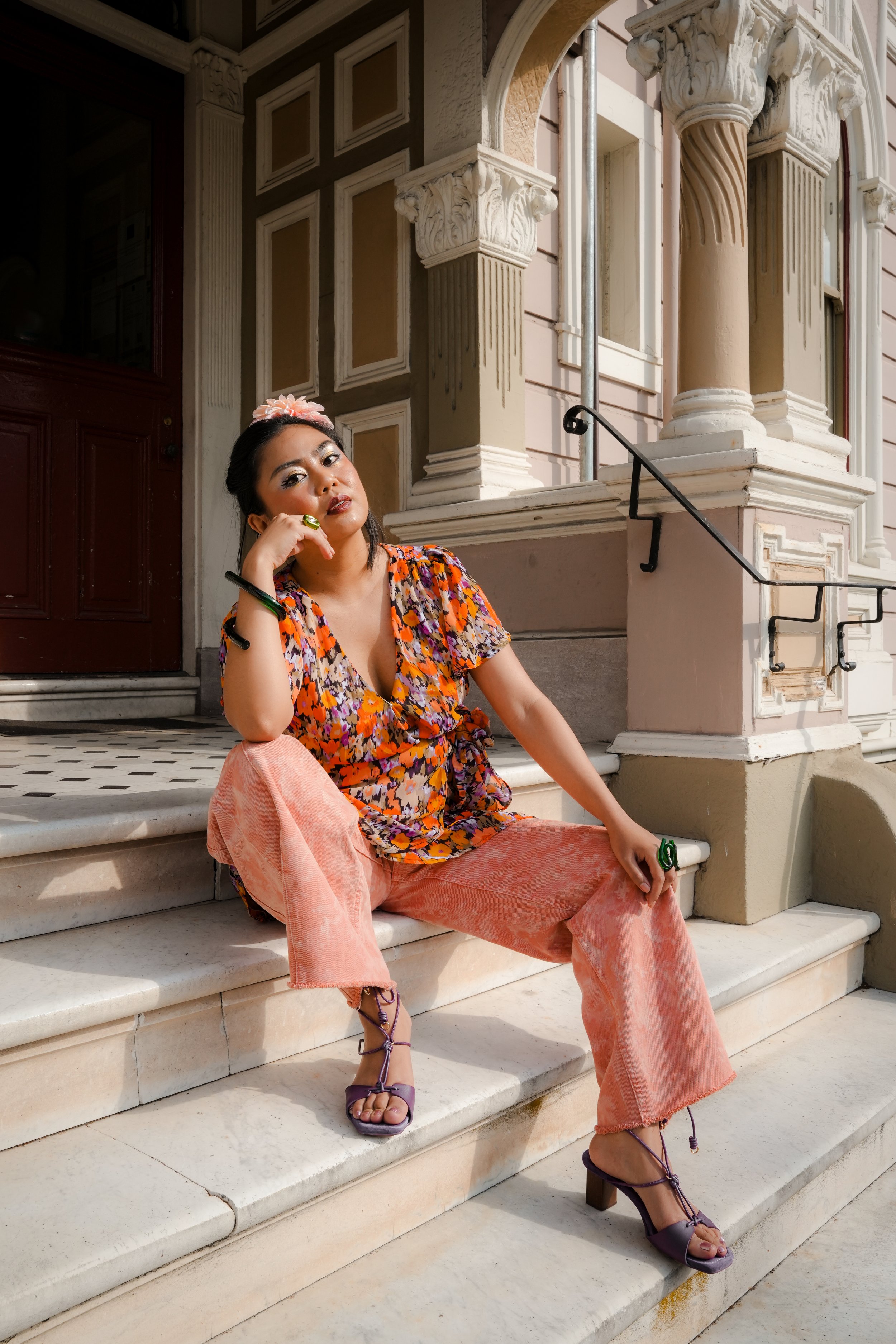 A woman with dark hair and makeup sitting on marble stairs outside a historic building, wearing a colorful floral top, peach pants, and purple high-heeled sandals, with her hand resting on her face.