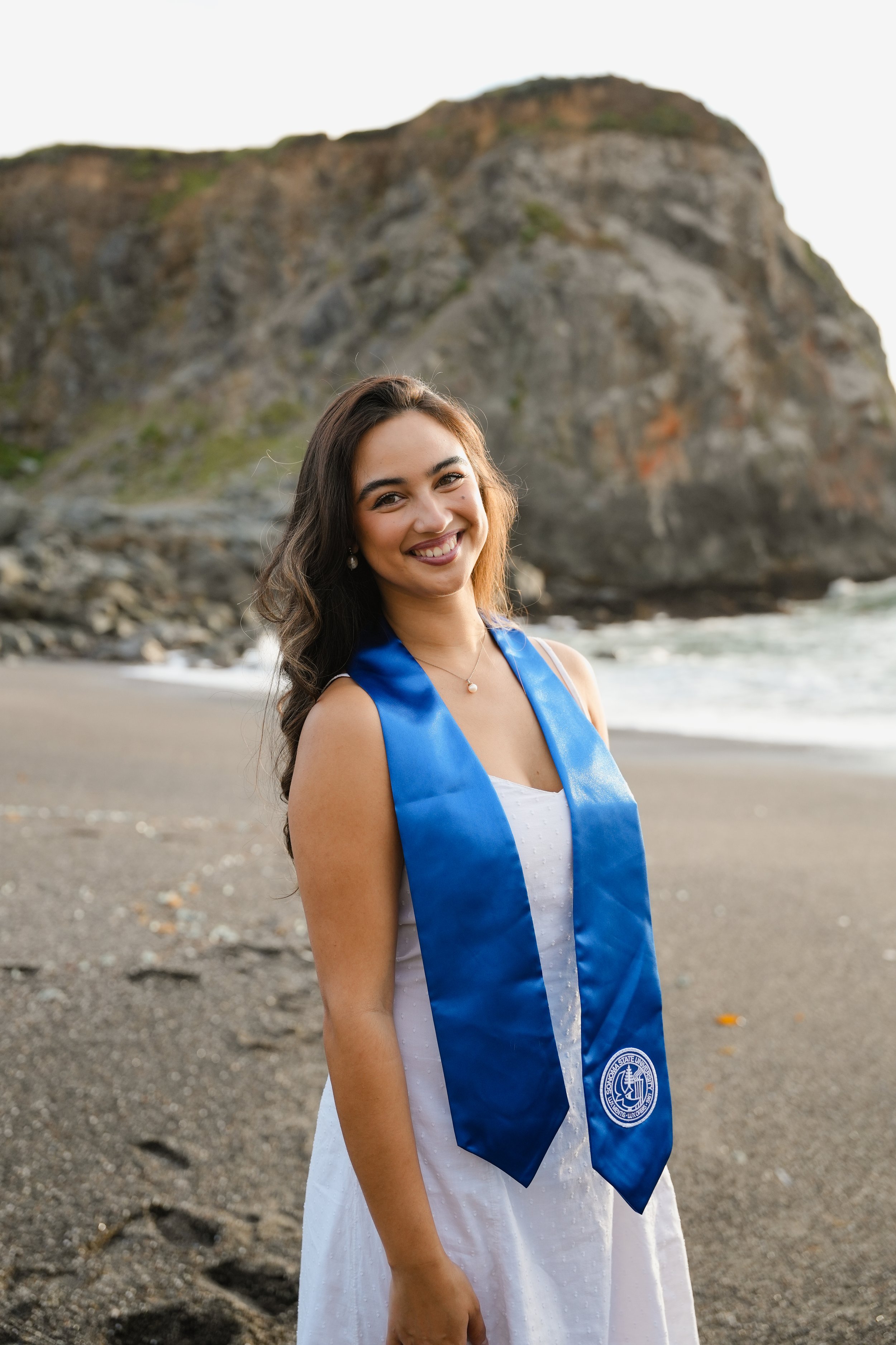 A young woman in a white dress and graduation stole standing on a rocky beach with a large cliff behind her, smiling at the camera during daylight.