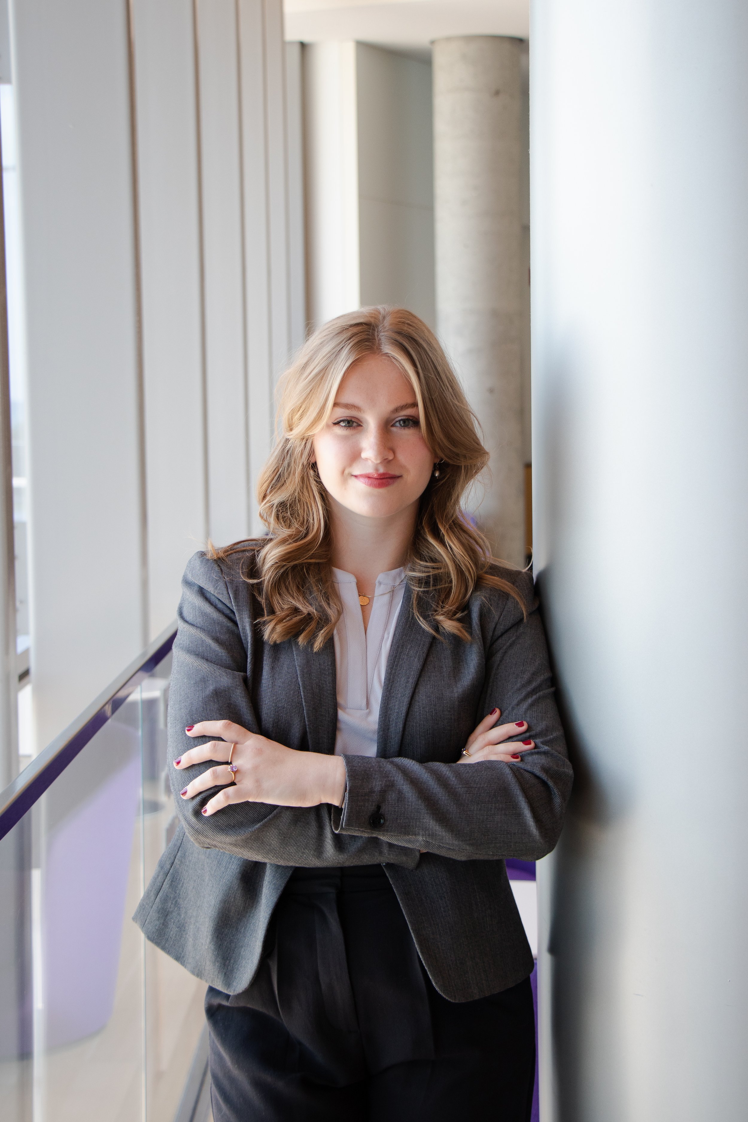 A professional woman with blonde wavy hair and light makeup standing with arms crossed, leaning against a white wall in a modern office building with large windows.