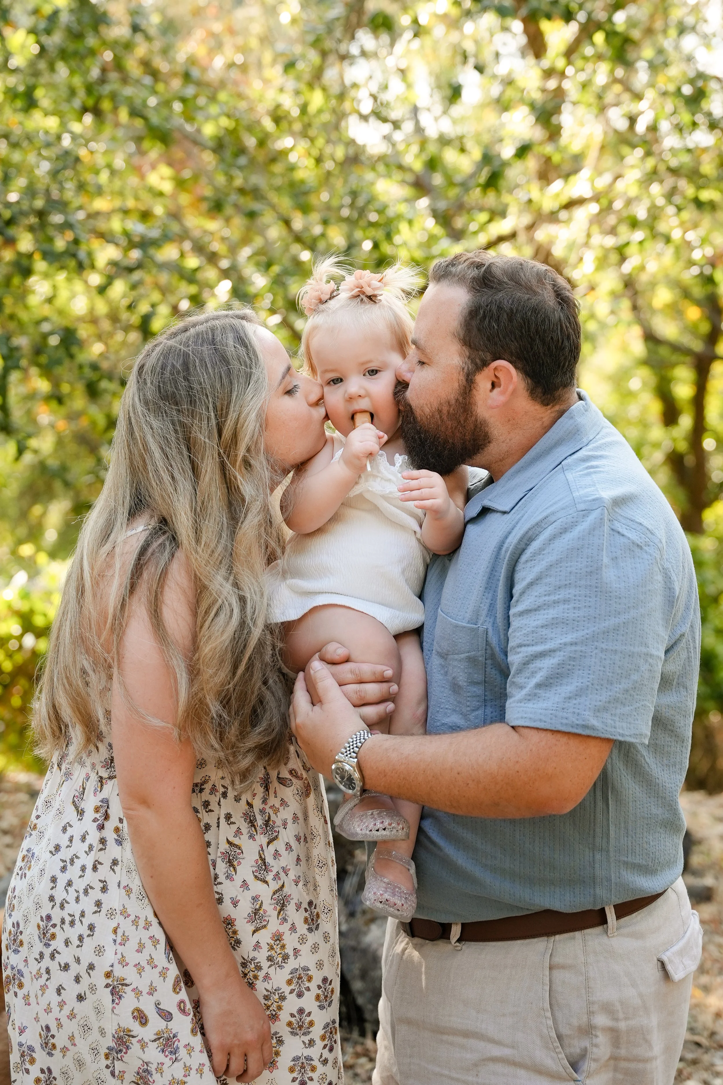 A family of three, including a woman, a man, and a young girl, sharing kisses and a hug outdoors in a park with green trees in the background.