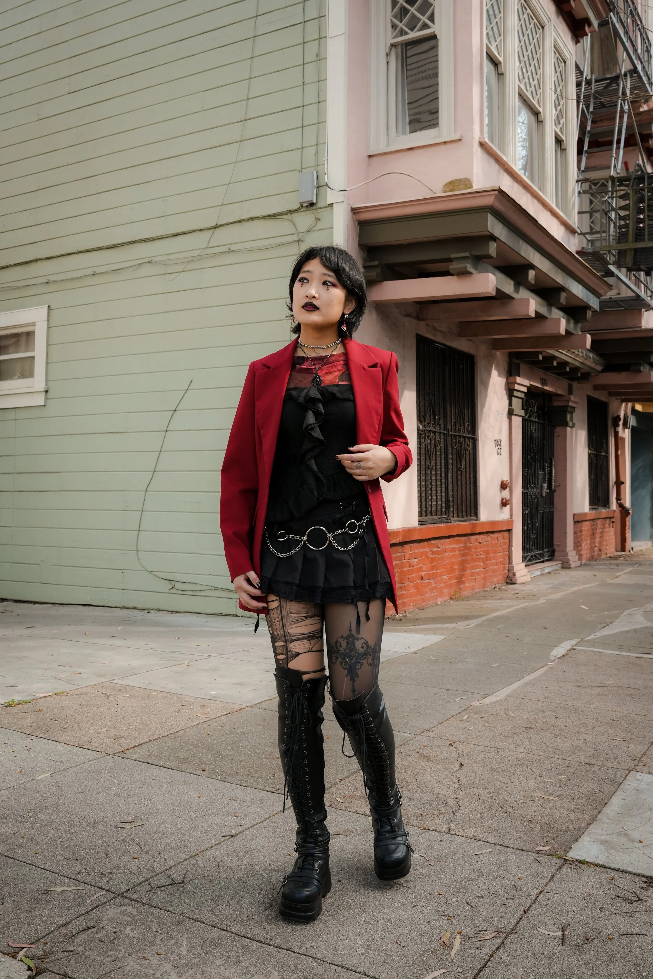 A young woman with black hair in a gothic style, wearing a red blazer, black dress with ruffles, and black ripped tights with a large black cross design. She has multiple necklaces, earrings, and dark makeup, including lipstick and eye makeup. She is