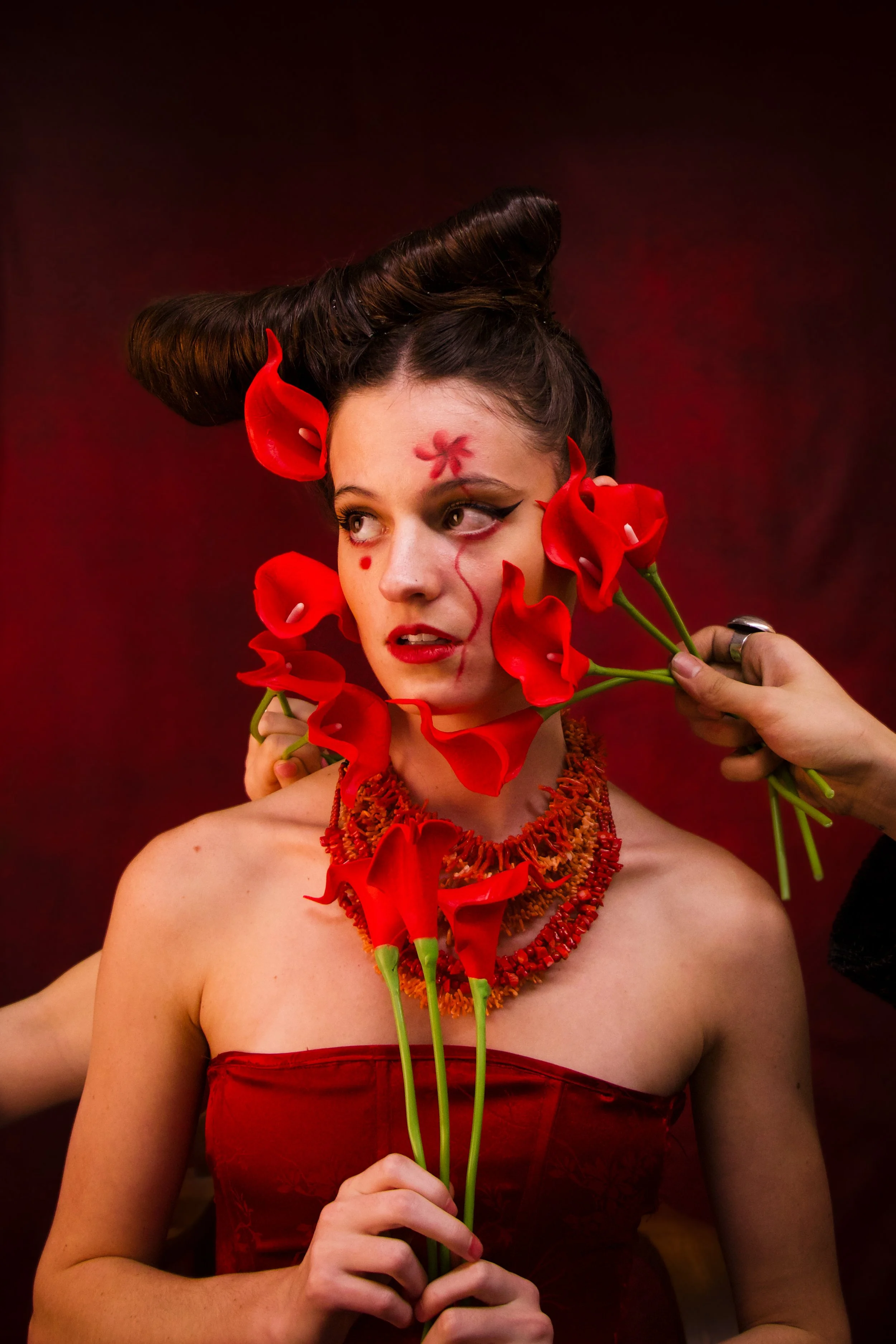 A woman dressed in a strapless red dress holding three red calla lilies. She has dramatic makeup with winged eyeliner, a red flower painted on her forehead, and red dots on her cheek. Her hair is styled in an elaborate updo with a large rolled sectio
