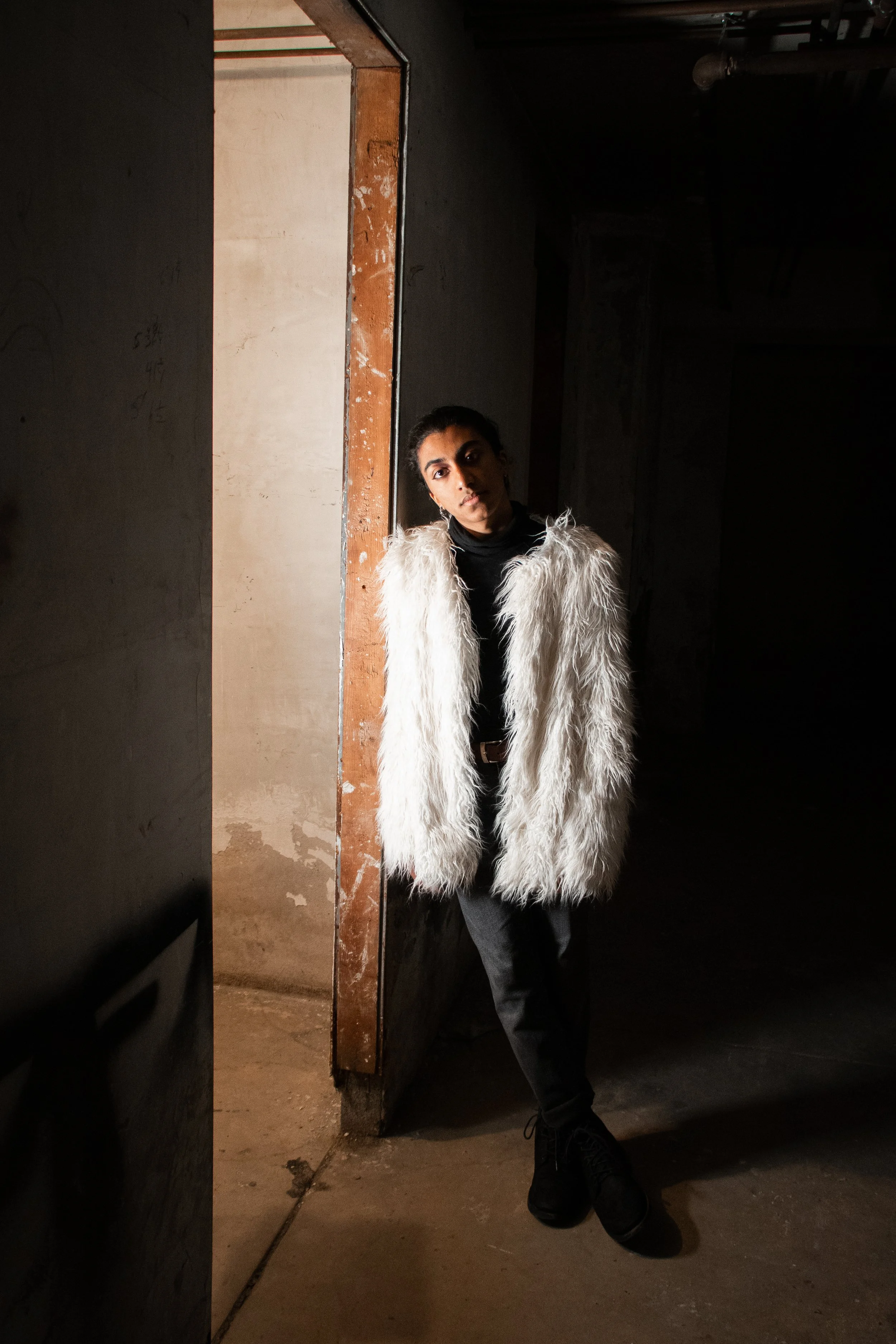 A woman with dark hair and a serious expression stands in a dimly lit space, wearing a white furry coat over a black top and black pants. She is leaning against a wall near an open doorway with an unfinished interior.