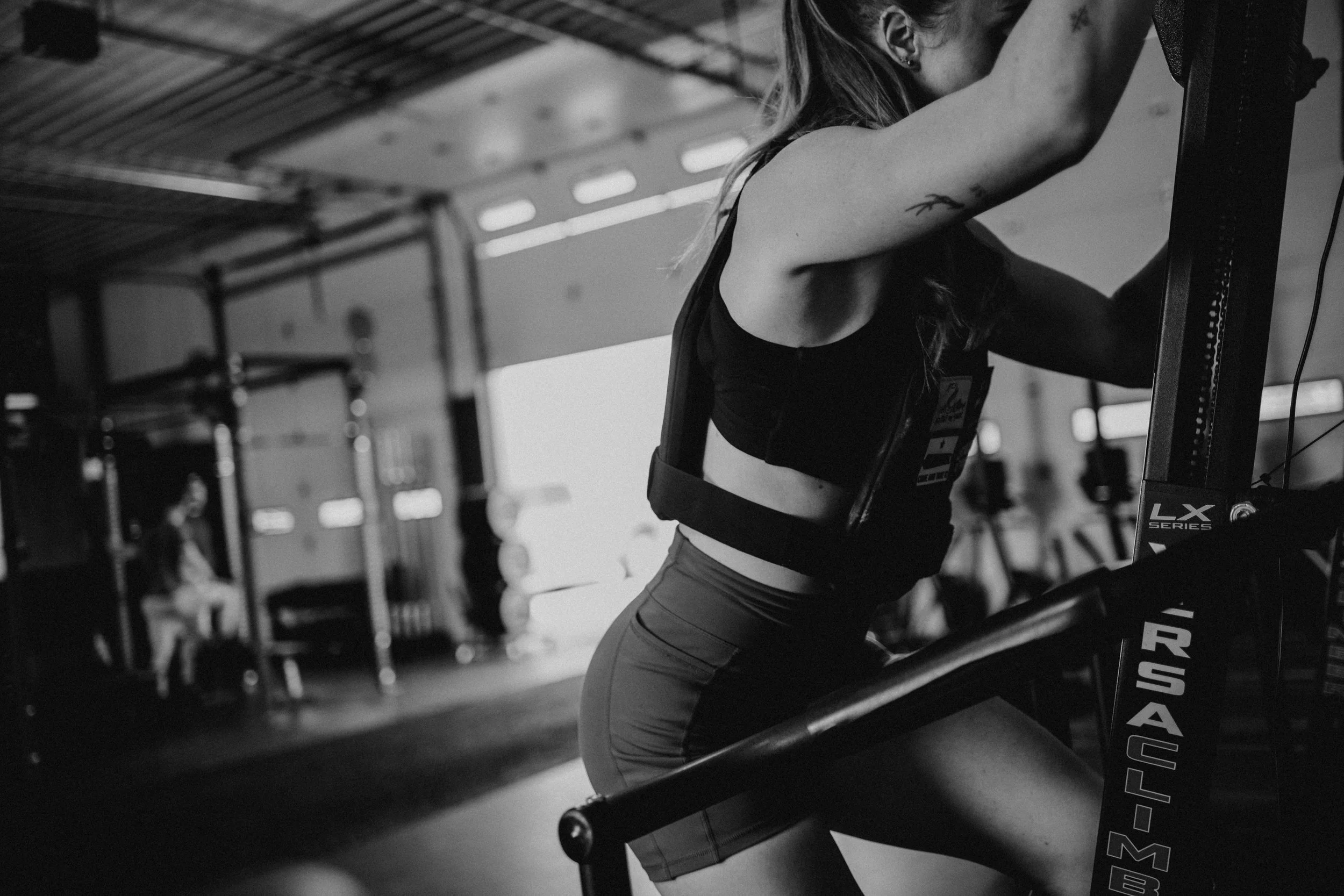 People working out in a gym with an open garage door, a man in the foreground lifting a dumbbell, and a sign that says 'No Parking Running Lane In Use!'