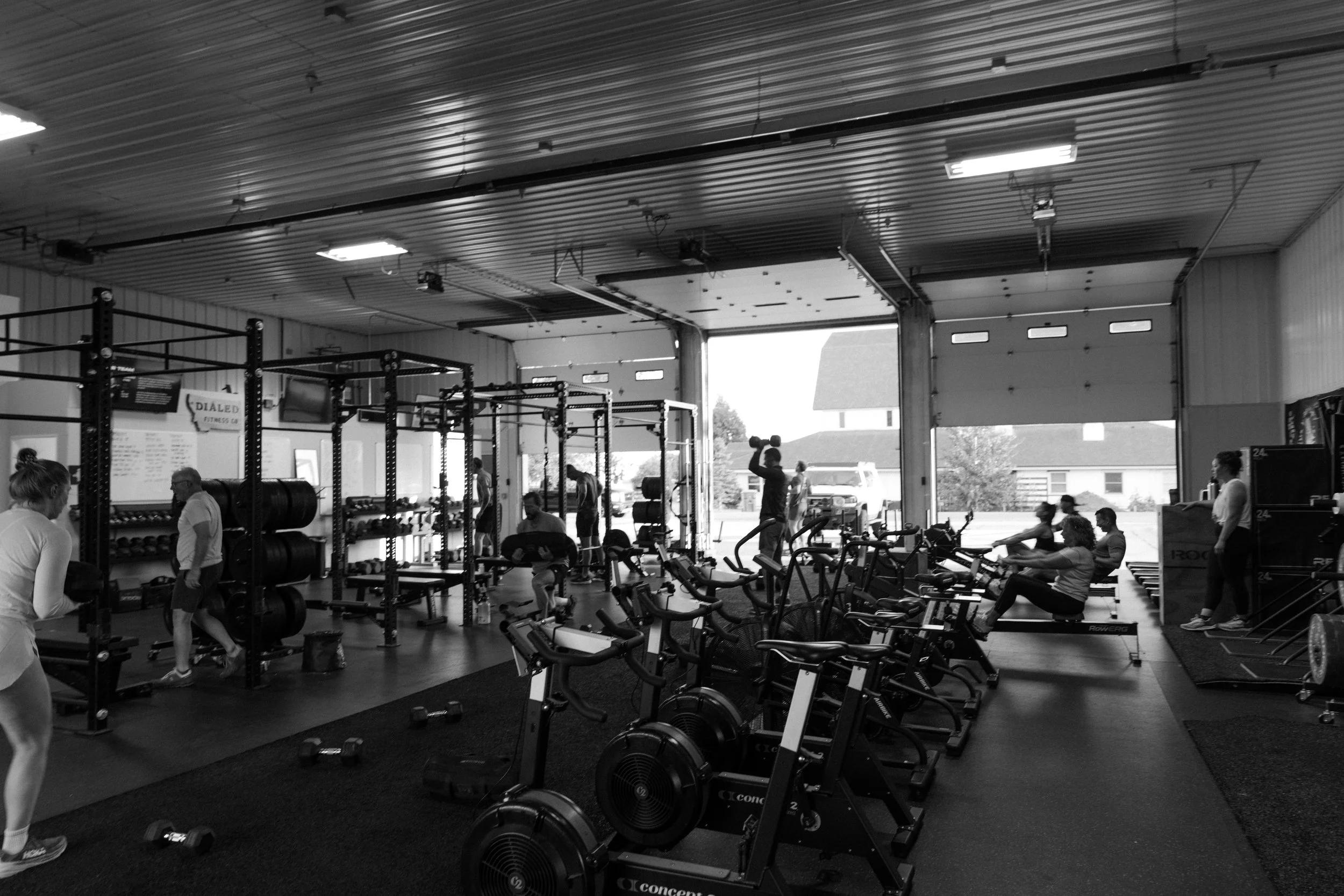 People working out in a gym with an open garage door, a man in the foreground lifting a dumbbell, and a sign that says 'No Parking Running Lane In Use!'