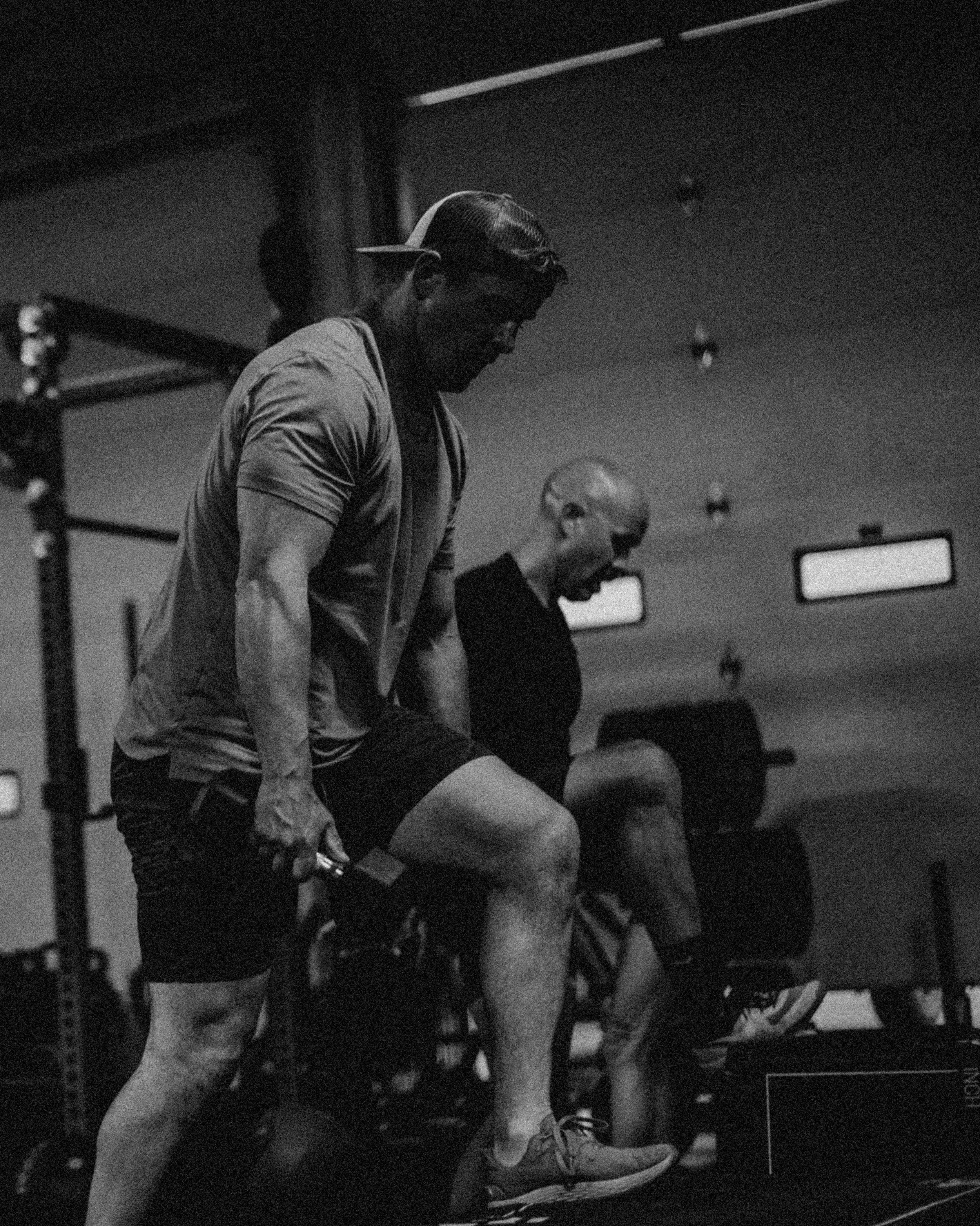 People working out in a gym with an open garage door, a man in the foreground lifting a dumbbell, and a sign that says 'No Parking Running Lane In Use!'