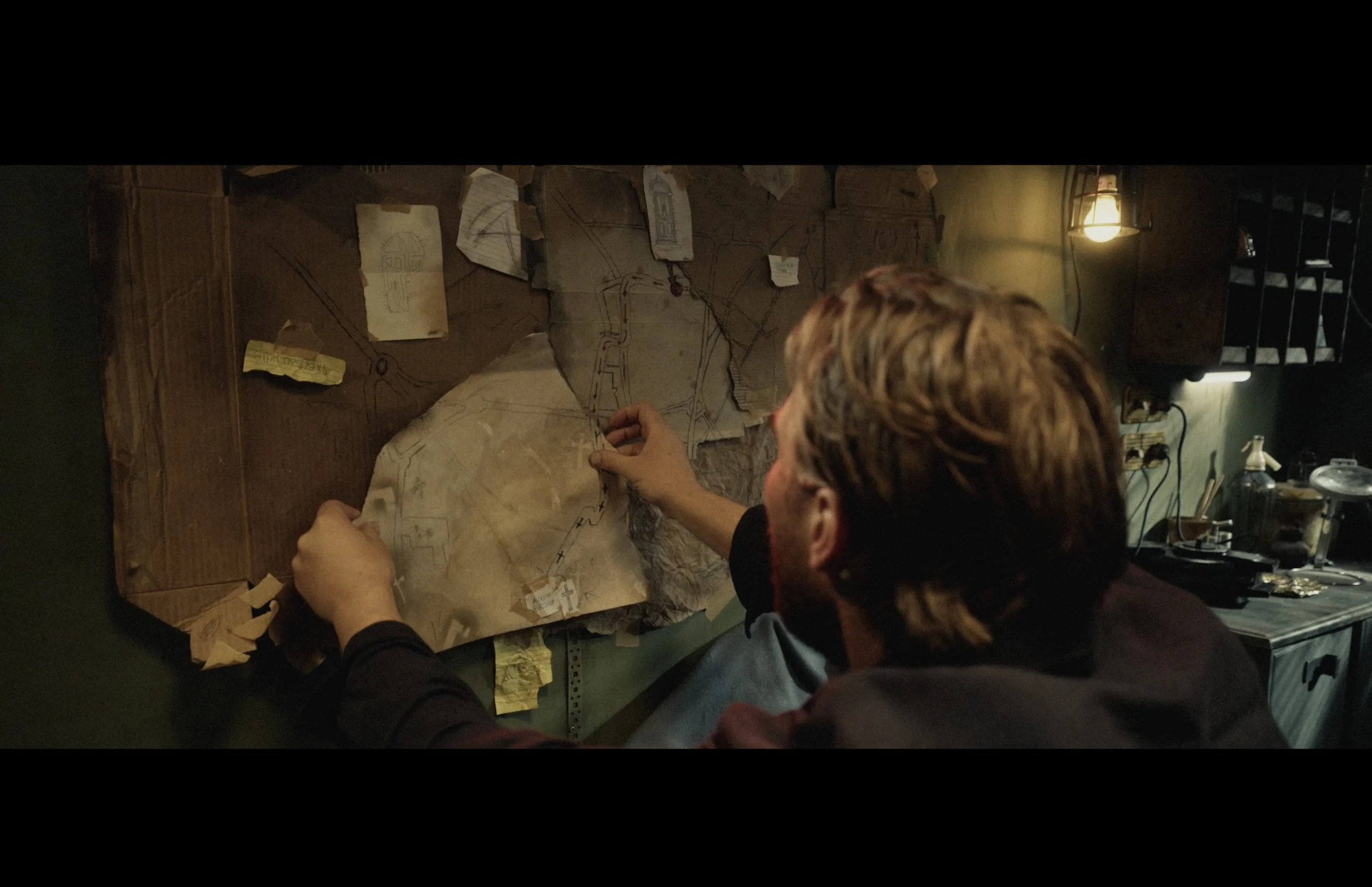 Person assembling a detailed map or blueprint on a cluttered desk, with papers and notes taped to a wooden wall behind.