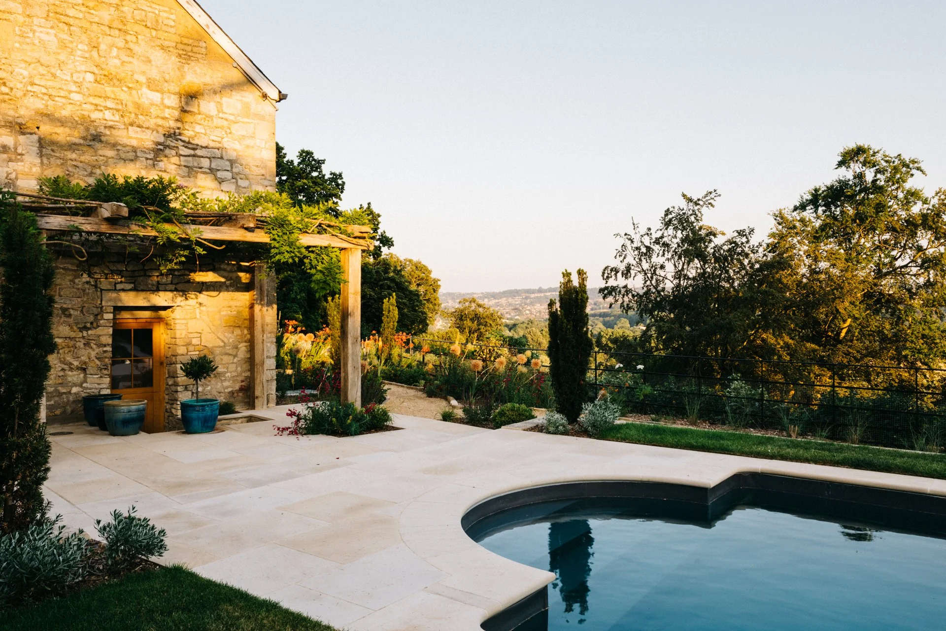 A garden with a swimming pool, stone house, outdoor plants, trees, and a view of the distant landscape in the evening sunlight.