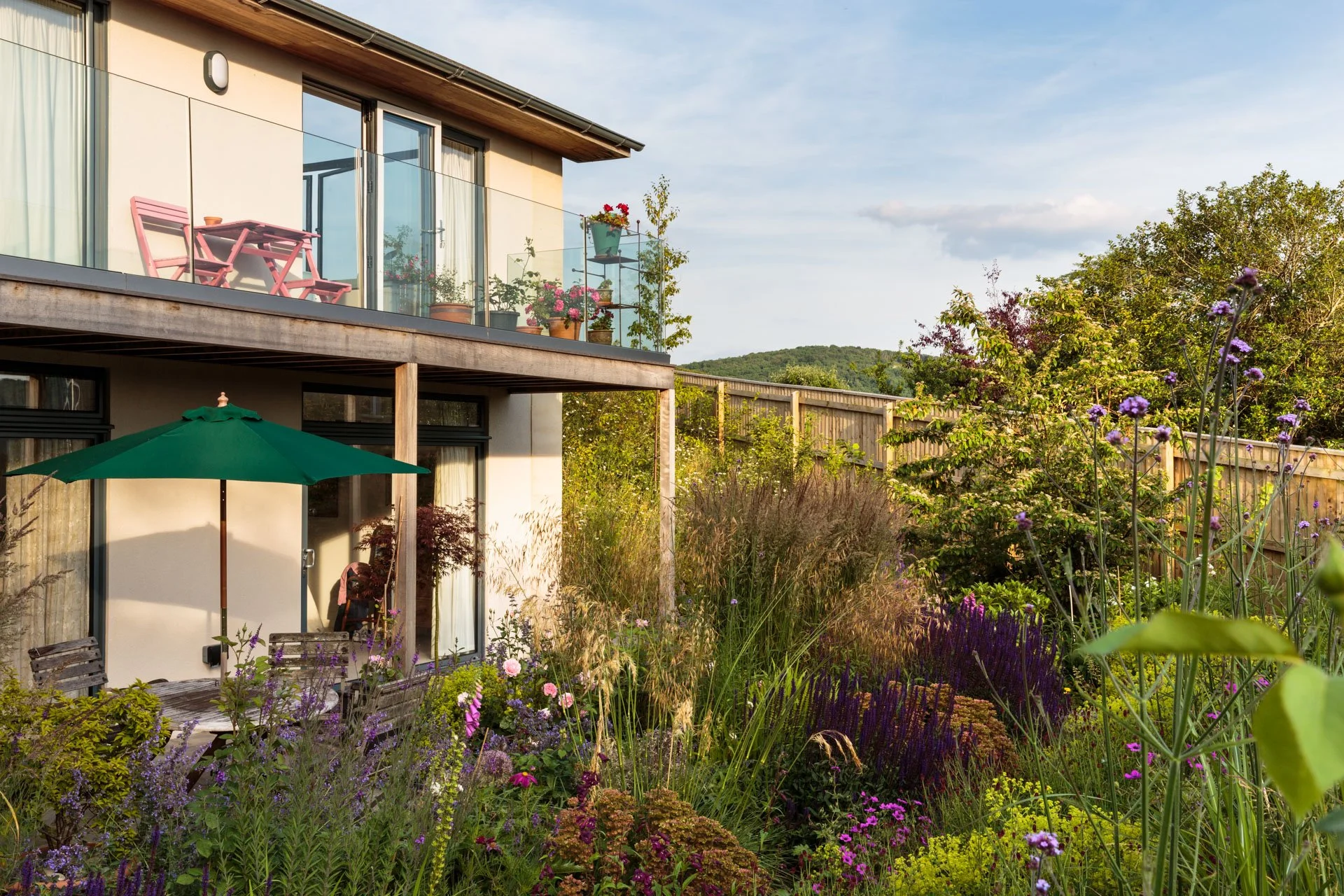 View of a modern house with a balcony decorated with potted plants and pink outdoor furniture, surrounded by a lush garden with various flowering plants, tall grasses, and trees, under a partly cloudy sky during daytime.