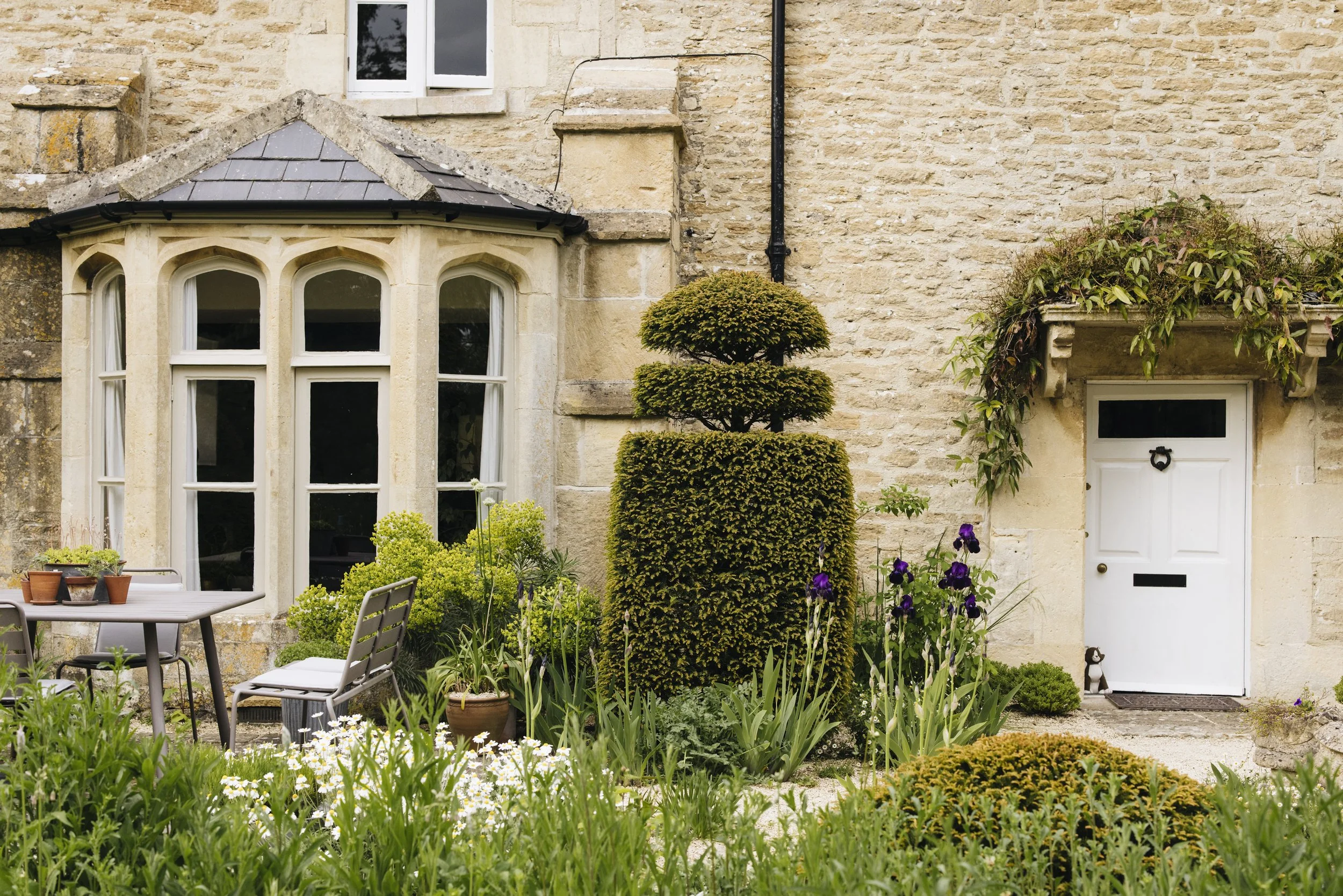 Front view of a stone house with a small garden and potted plants, featuring a bay window and a white door surrounded by lush greenery and flowering plants.