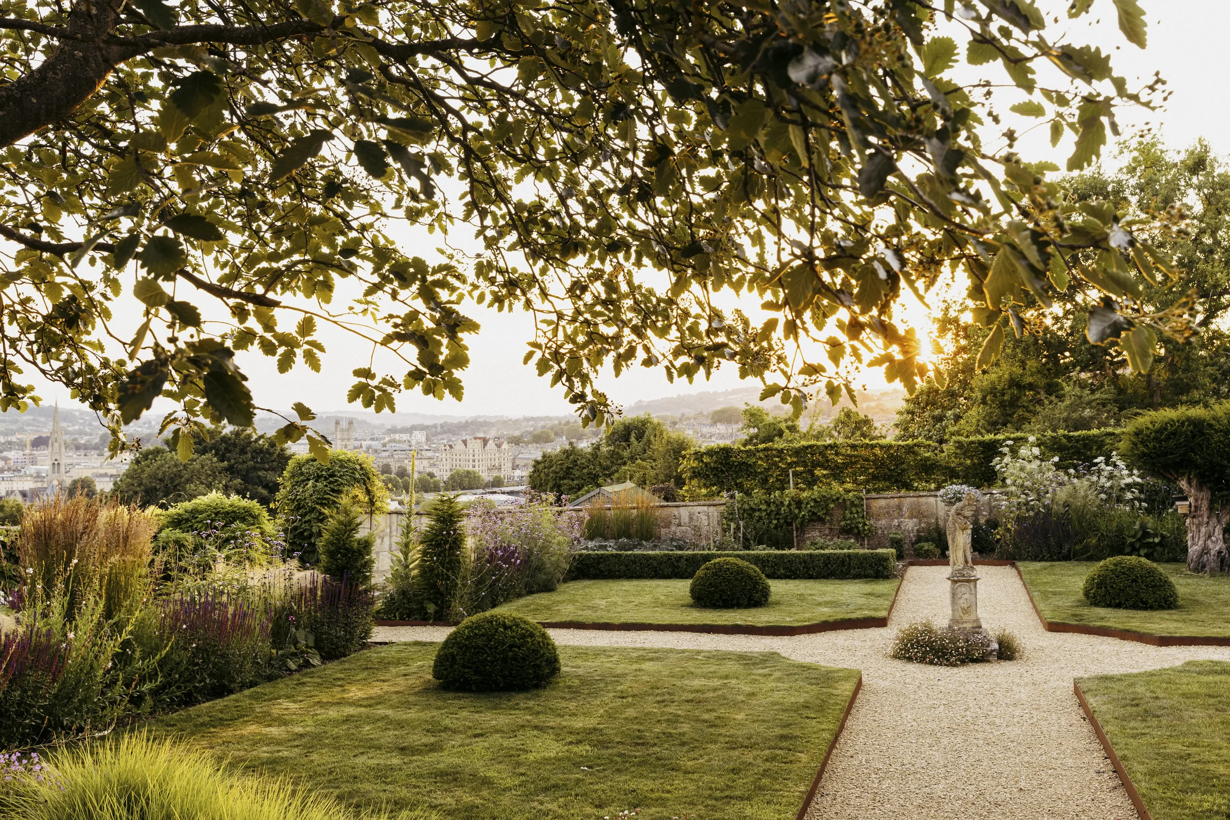 A garden with trimmed bushes, colorful flowers, a stone statue, and gravel pathways, with cityscape and trees in the background during sunset.