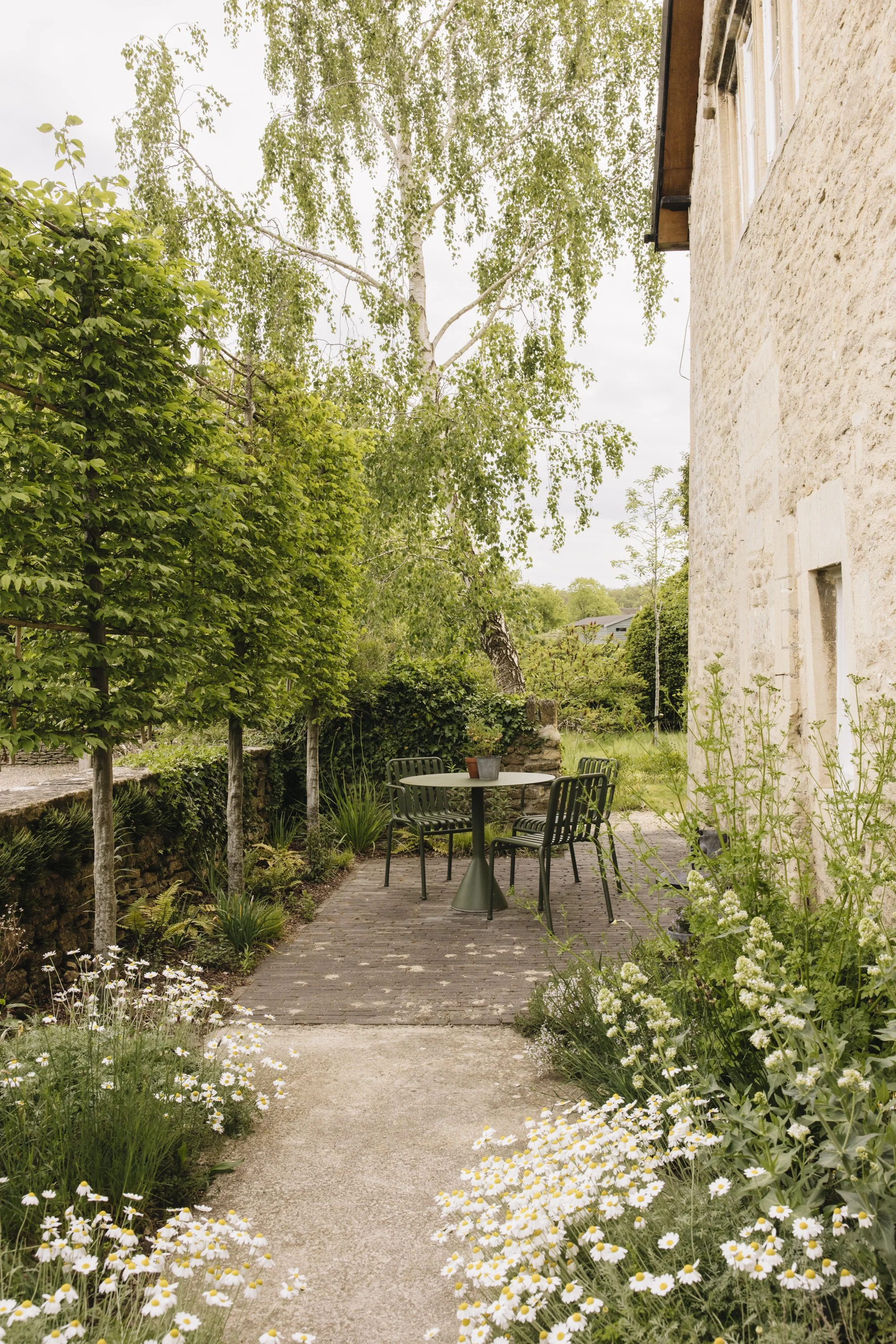 A charming outdoor garden patio with a round table and four black chairs, surrounded by lush green plants and white flowering bushes, next to a rustic stone building with white window frames.