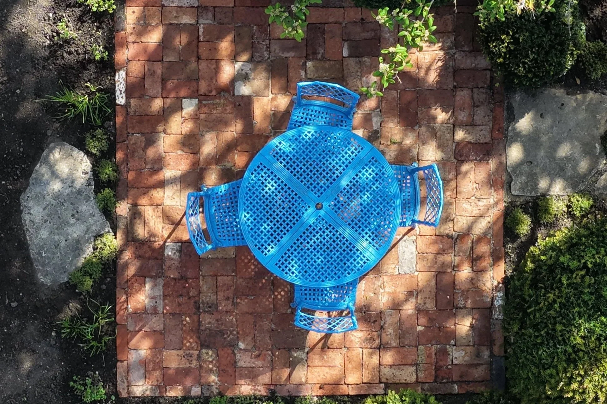 Blue metal patio table and two matching chairs on a brick patio, surrounded by small plants and greenery. Shadows cast on the brick surface.