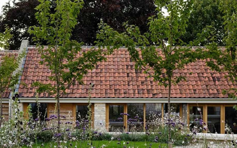 A house with a red tiled roof, wooden window frames, surrounded by green trees and a garden with various flowering plants.