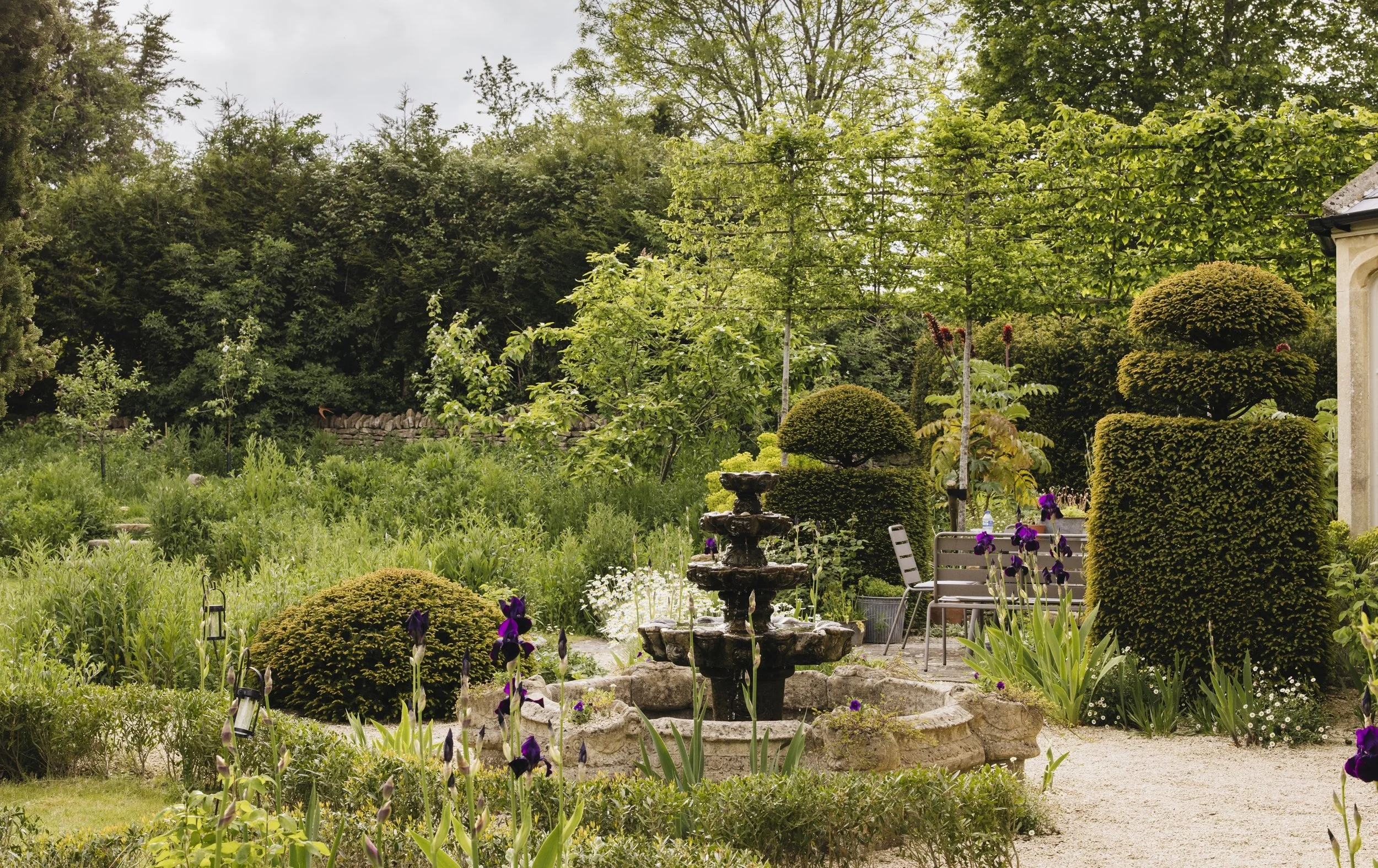 A lush garden with various trees, shrubs, and flowering plants, including purple irises. A stone fountain with water is centered in the foreground. There's a seating area with chairs, and a stone building wall visible on the right. The background shows dense green foliage and tall trees under an overcast sky.