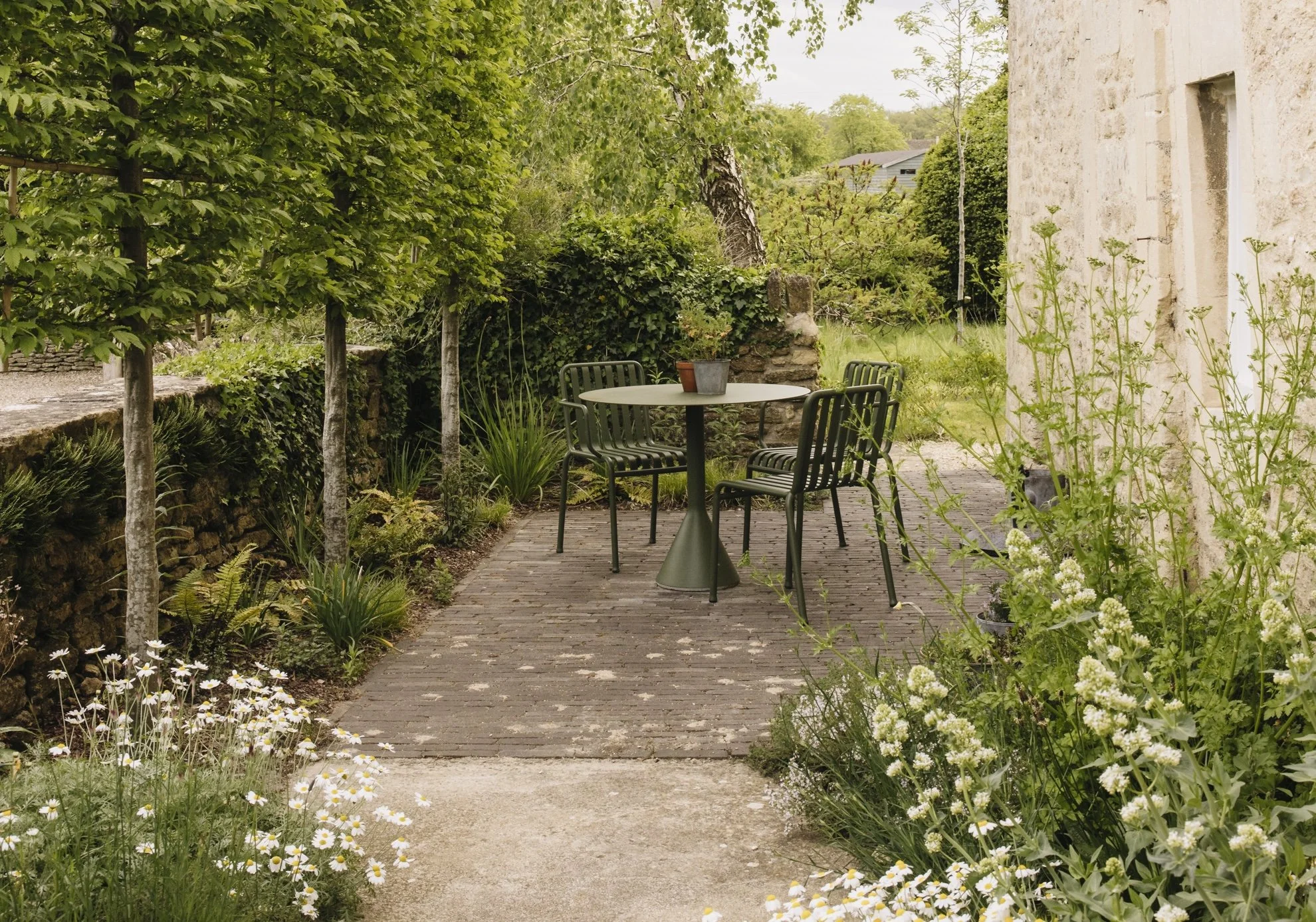 A garden patio with a round white table and four black chairs, surrounded by lush green trees and blooming flowers, next to a beige stone building.