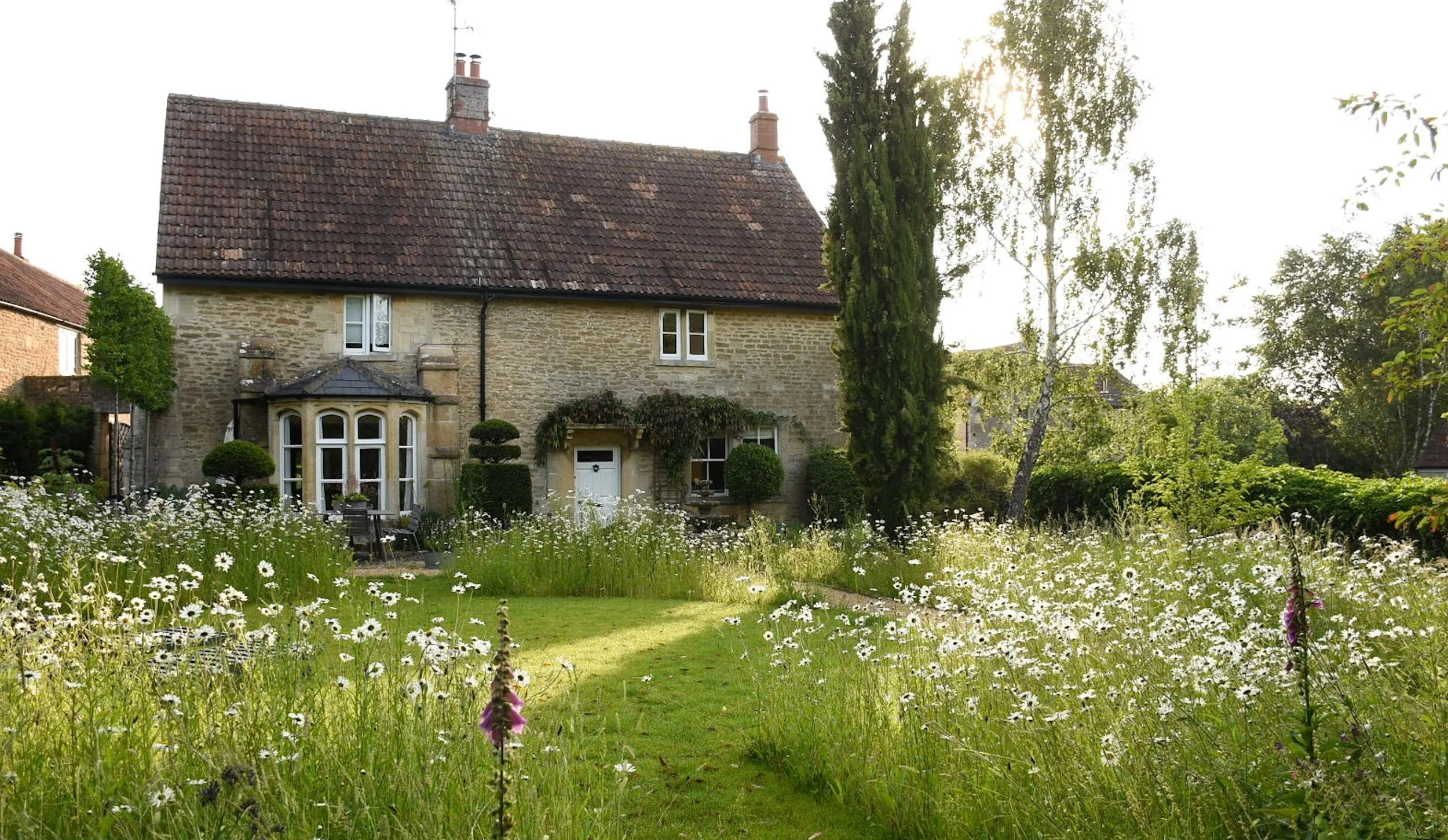 A stone house with a red tile roof, surrounded by a garden with daisies and trees, with sunlight filtering through the leaves.
