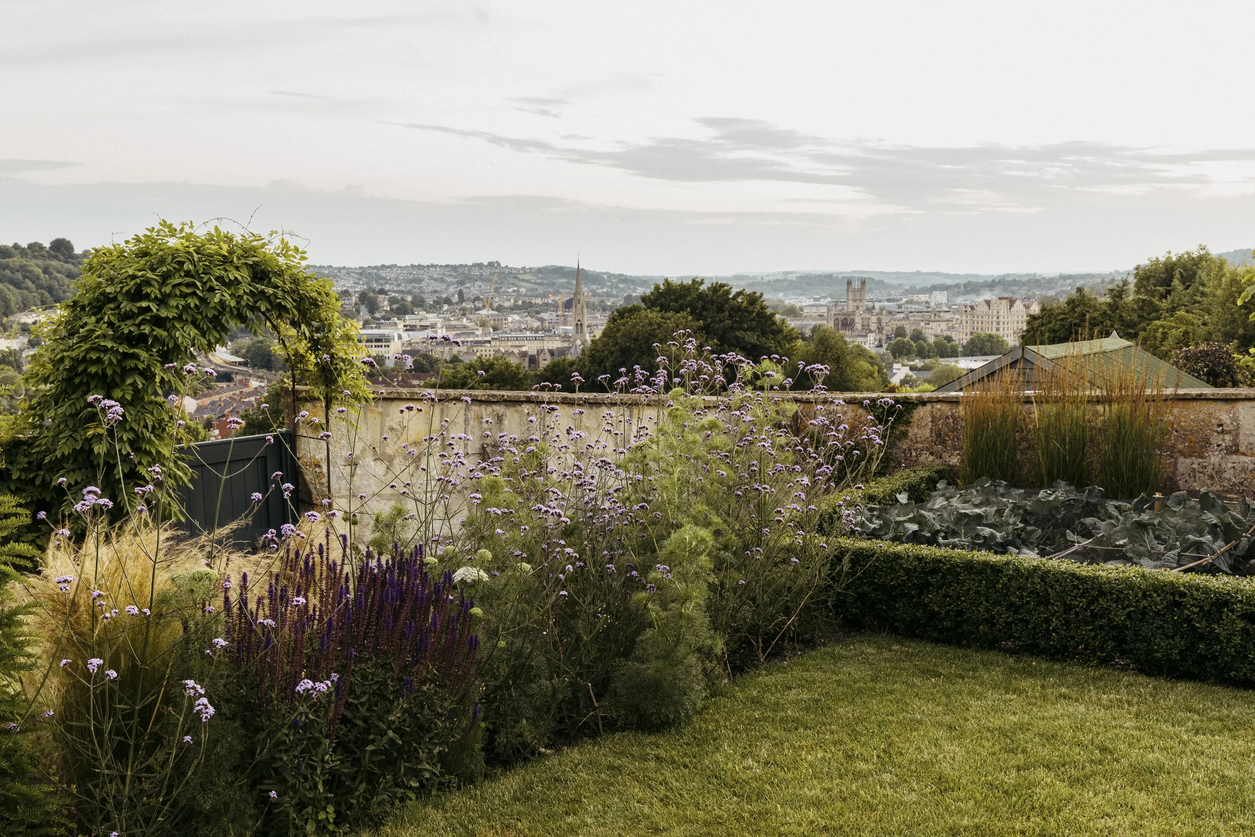 View of a city skyline from a garden with flowering plants, trees, and a stone wall in the foreground.