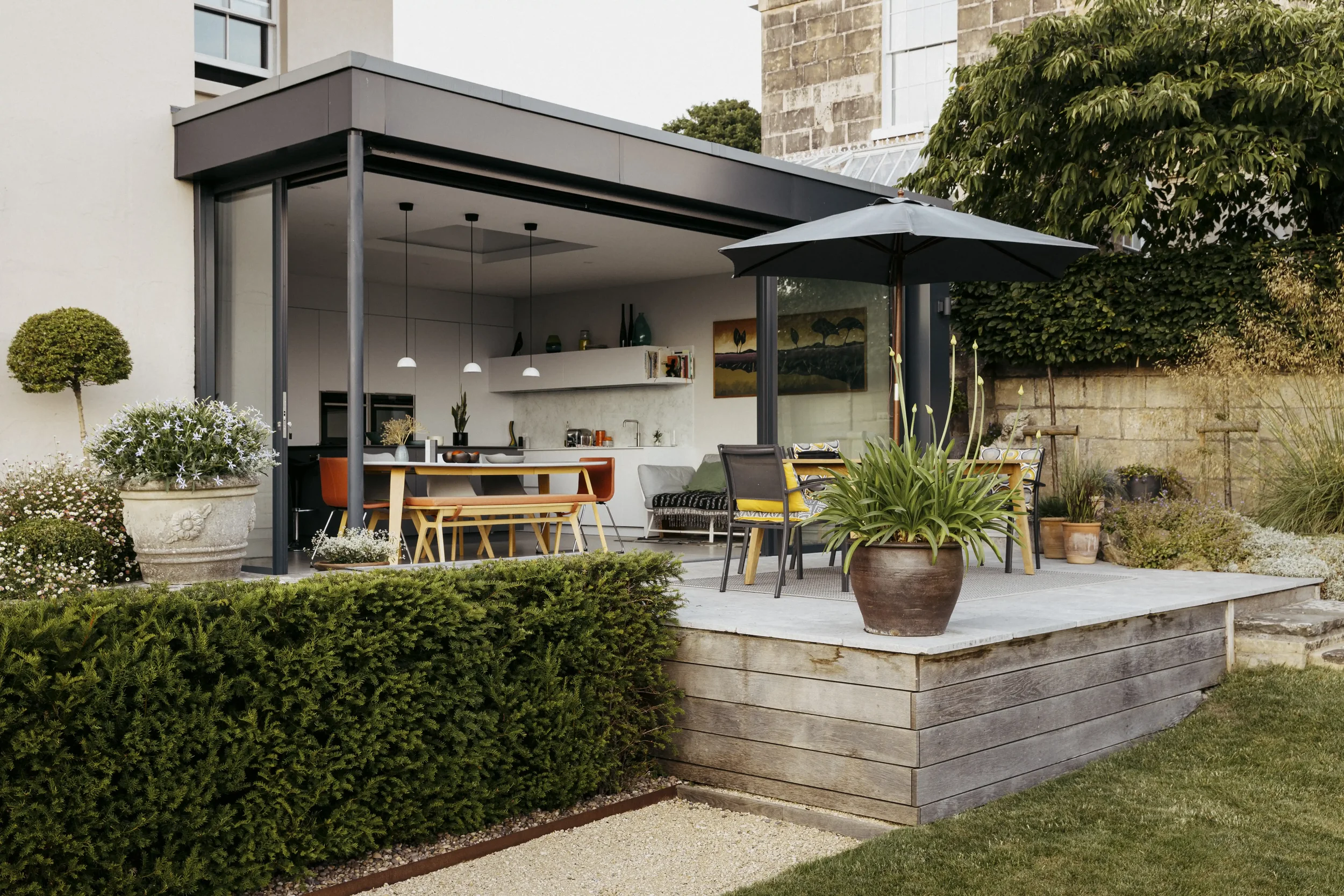 A modern outdoor patio with a built-in kitchen, dining area, and garden, featuring a black umbrella and potted plants.