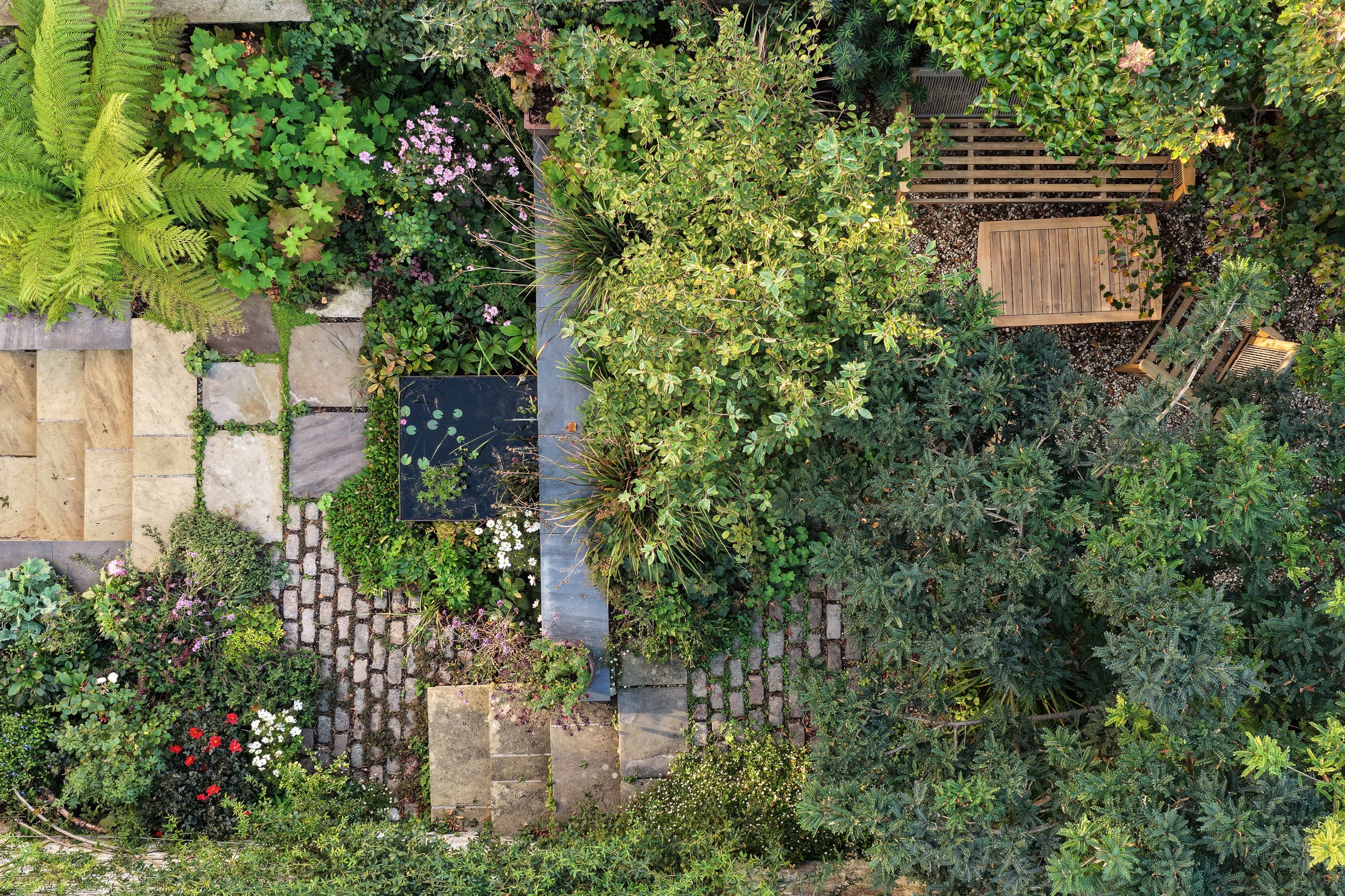 An aerial view of a lush backyard garden with a variety of green trees, flowering plants, stone pathways, and a wooden deck with chairs.
