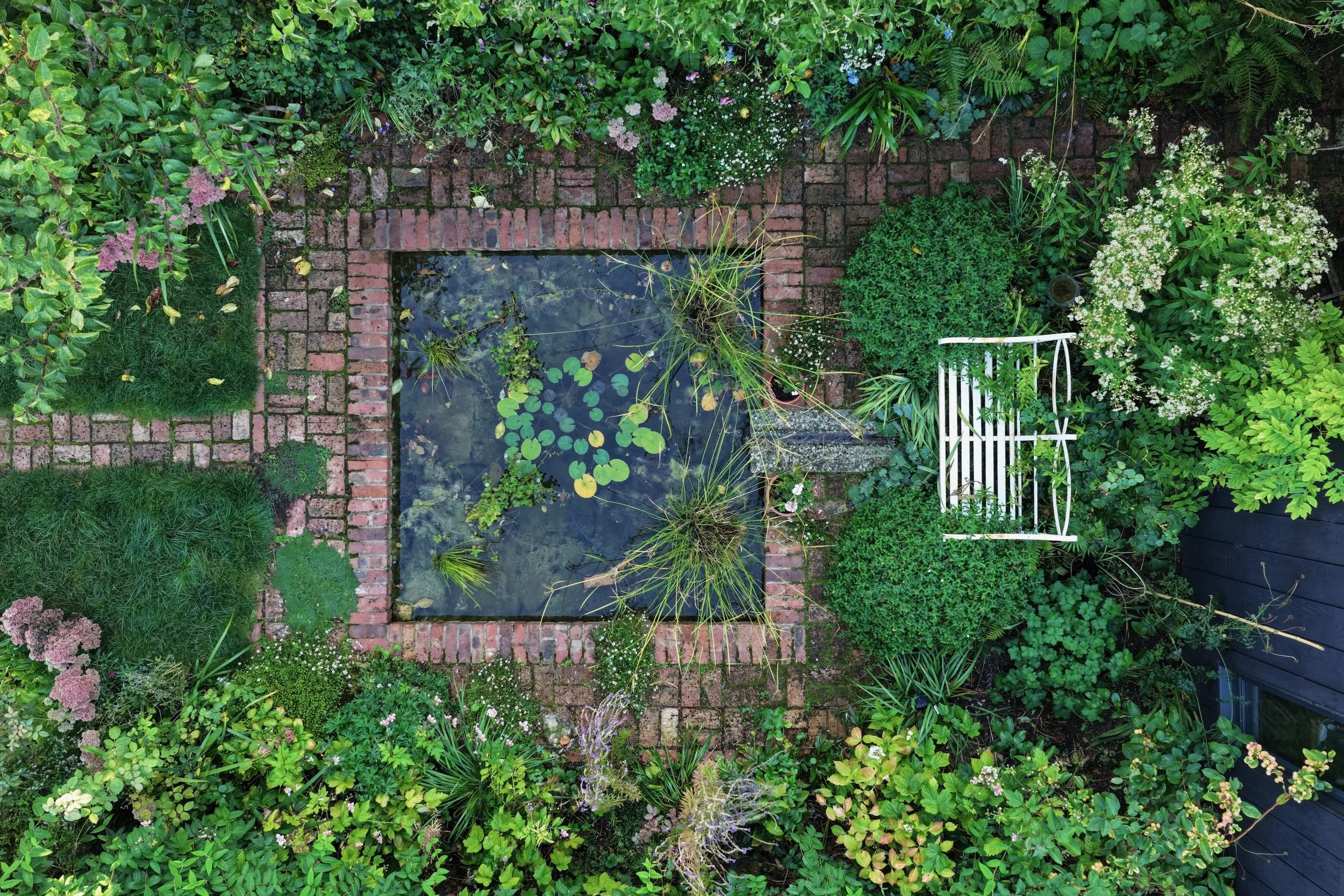 Top-down view of a lush garden with a small pond, brick border, stone path, and various green plants and flowers, including a white garden bench.