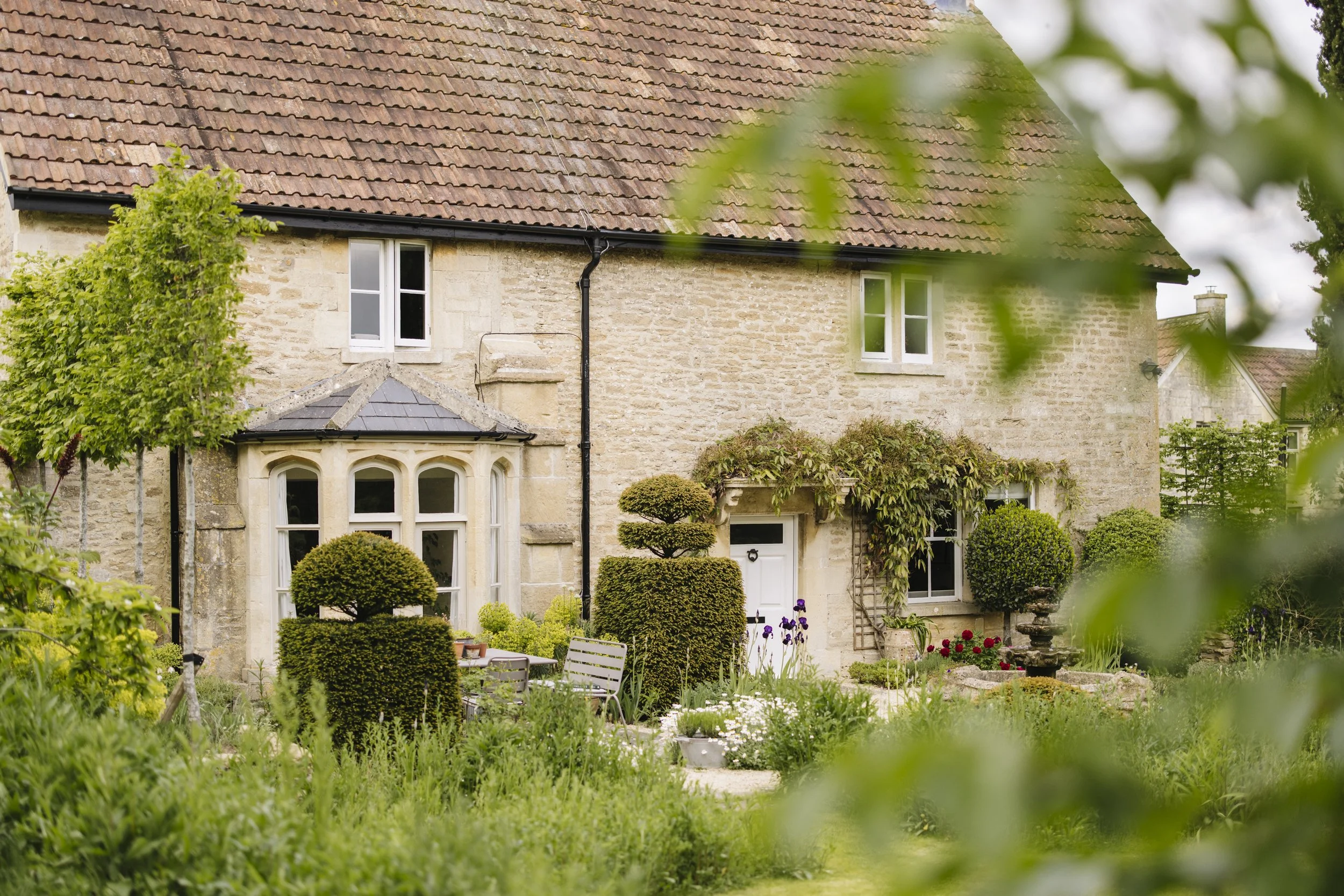 A stone house with a red tiled roof, surrounded by a lush green garden with trimmed bushes, flowers, and outdoor seating, partially framed by out-of-focus green leaves in the foreground.