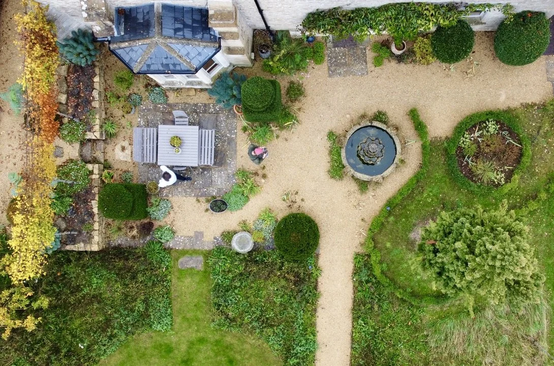 Aerial view of a spacious garden with a stone pathway, a round pond with a decorative fountain, lush green bushes, trees, and a patio area with a table and chairs, adjacent to a house with a blue roof.