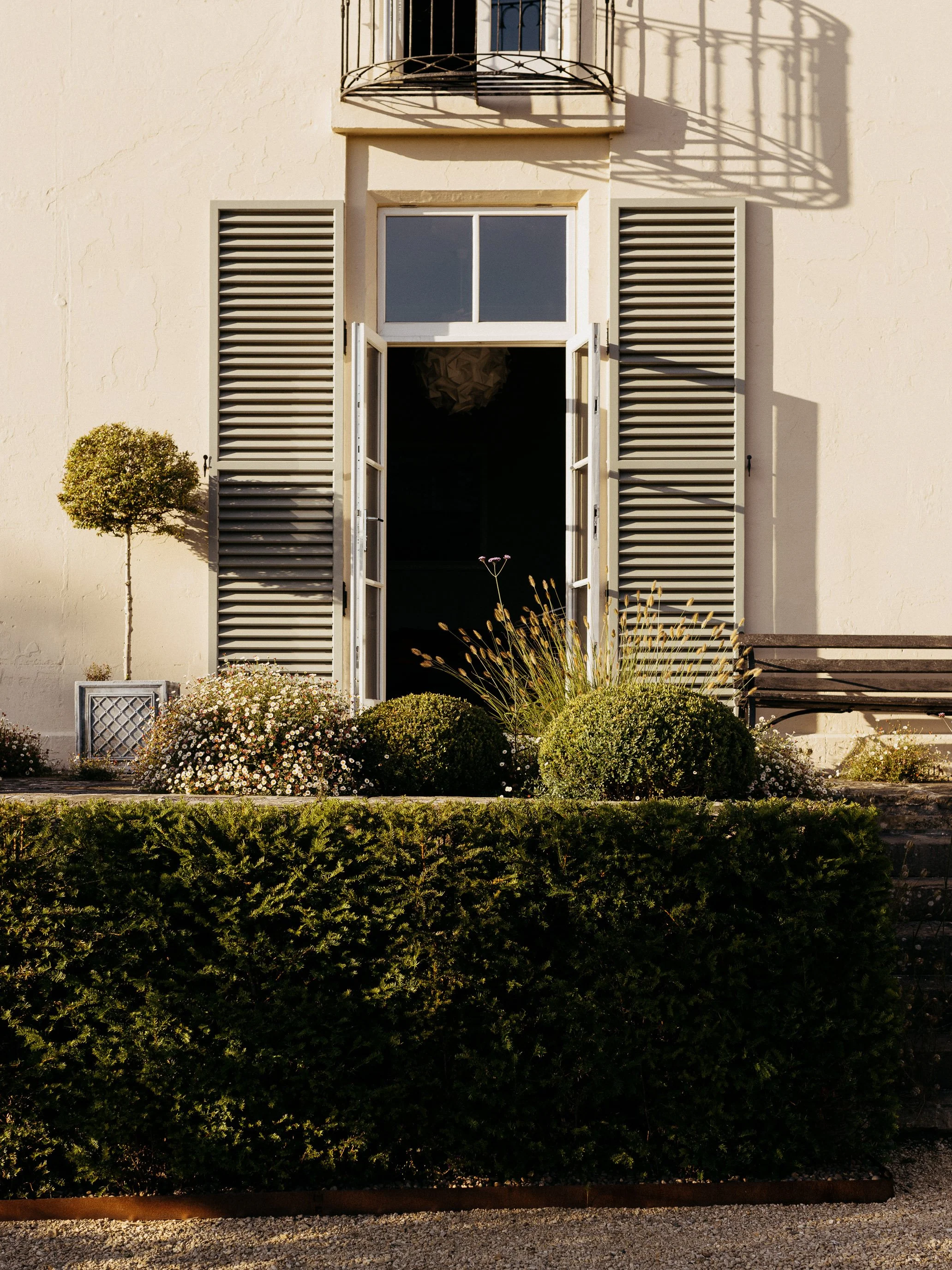 Close-up of a shuttered window and stone facade framed by clipped box balls and a timber bench