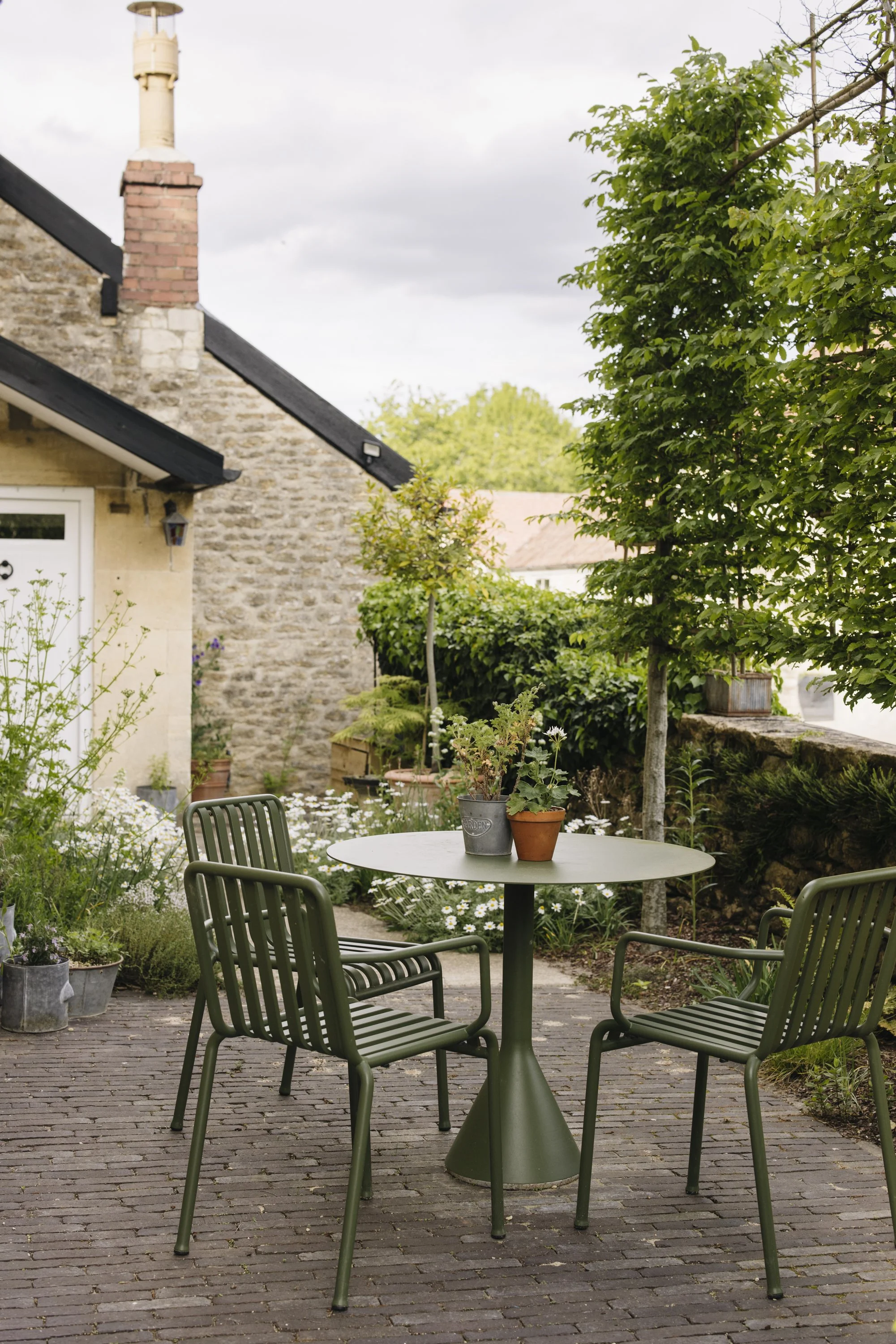 A round outdoor table with four dark green chairs on a brick patio. Two potted plants are on the table, and there are more plants and trees surrounding the patio. In the background, there is a house with stone and stucco walls, a brick chimney, and a sloped roof. The sky is cloudy.