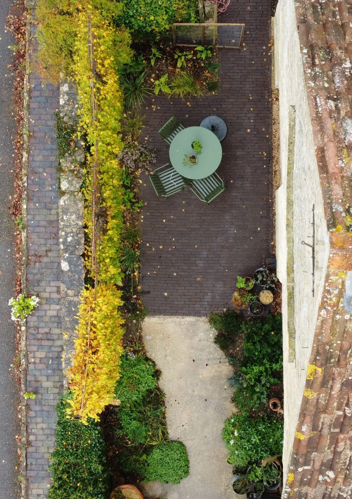 A small outdoor patio area viewed from above, with a round table, four chairs, potted plants, and a small garden with yellow and green foliage along the back wall.