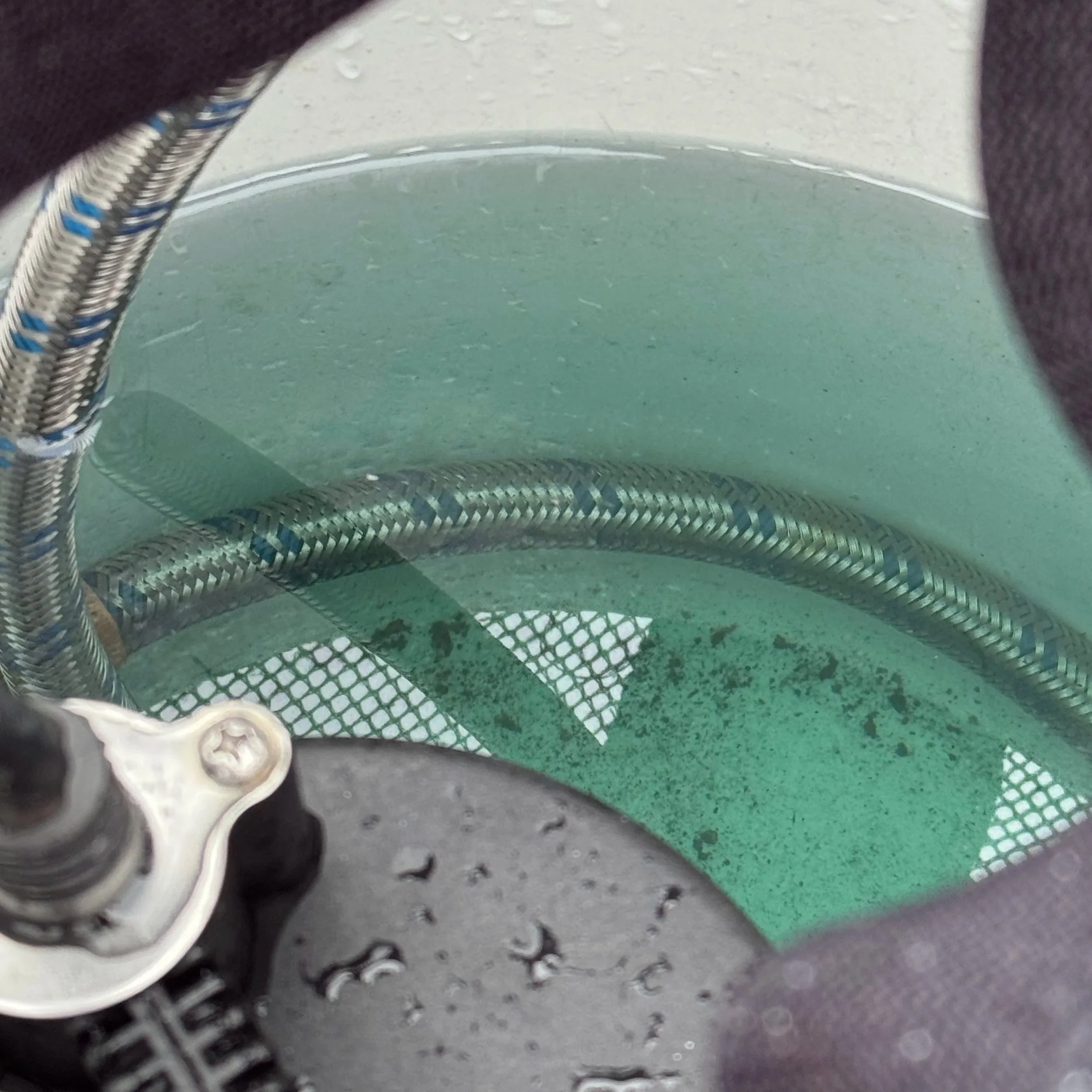 Looking down into a clear tank or container with a greenish tint, part of a metal hose inside, and some dirt or algae, with a black component and water droplets visible at the top edge.