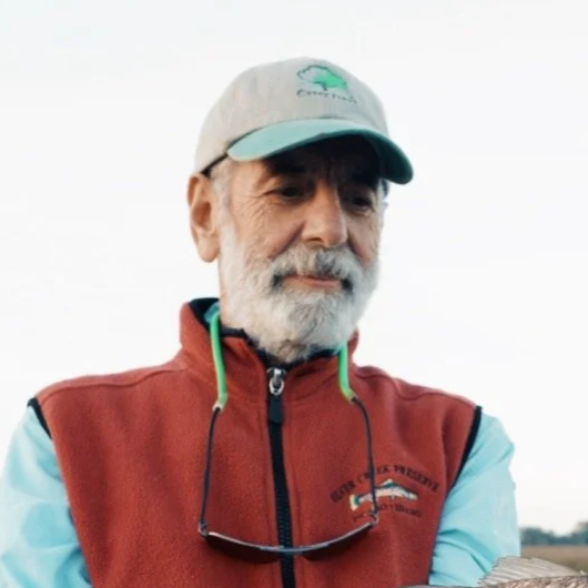 Close-up of an older man with gray hair and beard, wearing a gray and teal baseball cap, red vest, and sunglasses hanging around his neck.