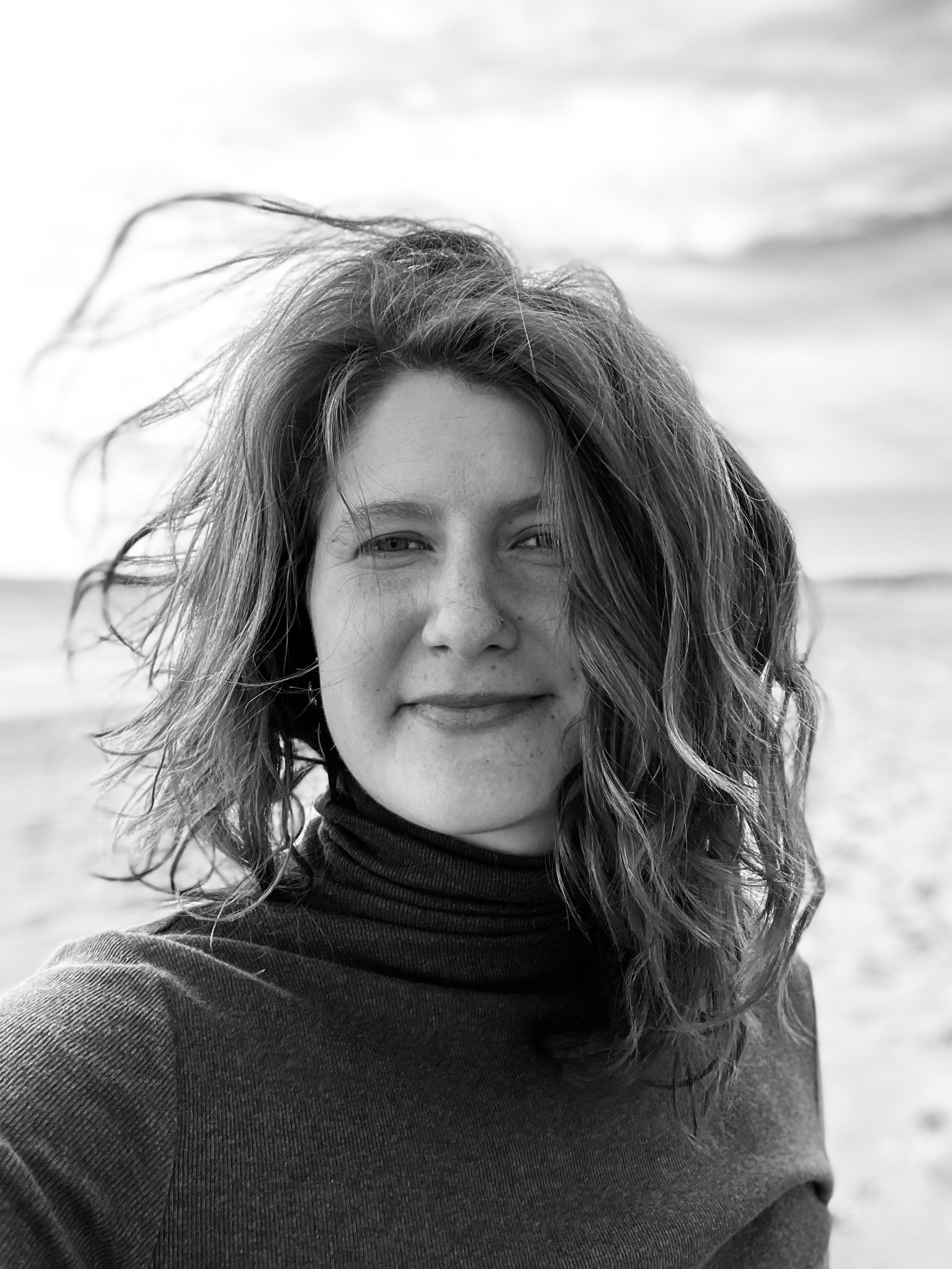 A woman with wavy hair smiling at the camera on a beach with the ocean in the background, black and white photo.