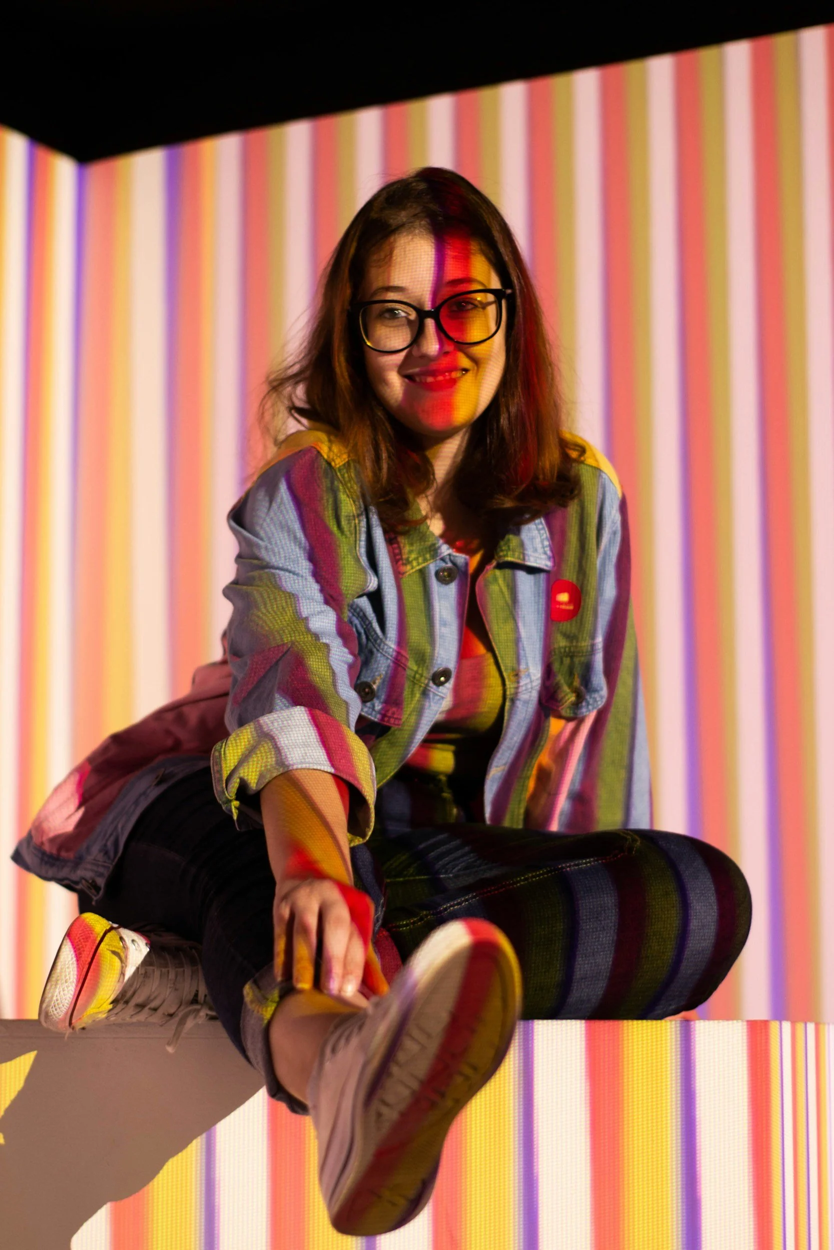 Young woman wearing glasses, sitting cross-legged on a platform, smiling, with colorful striped background and rainbow light reflections on her face