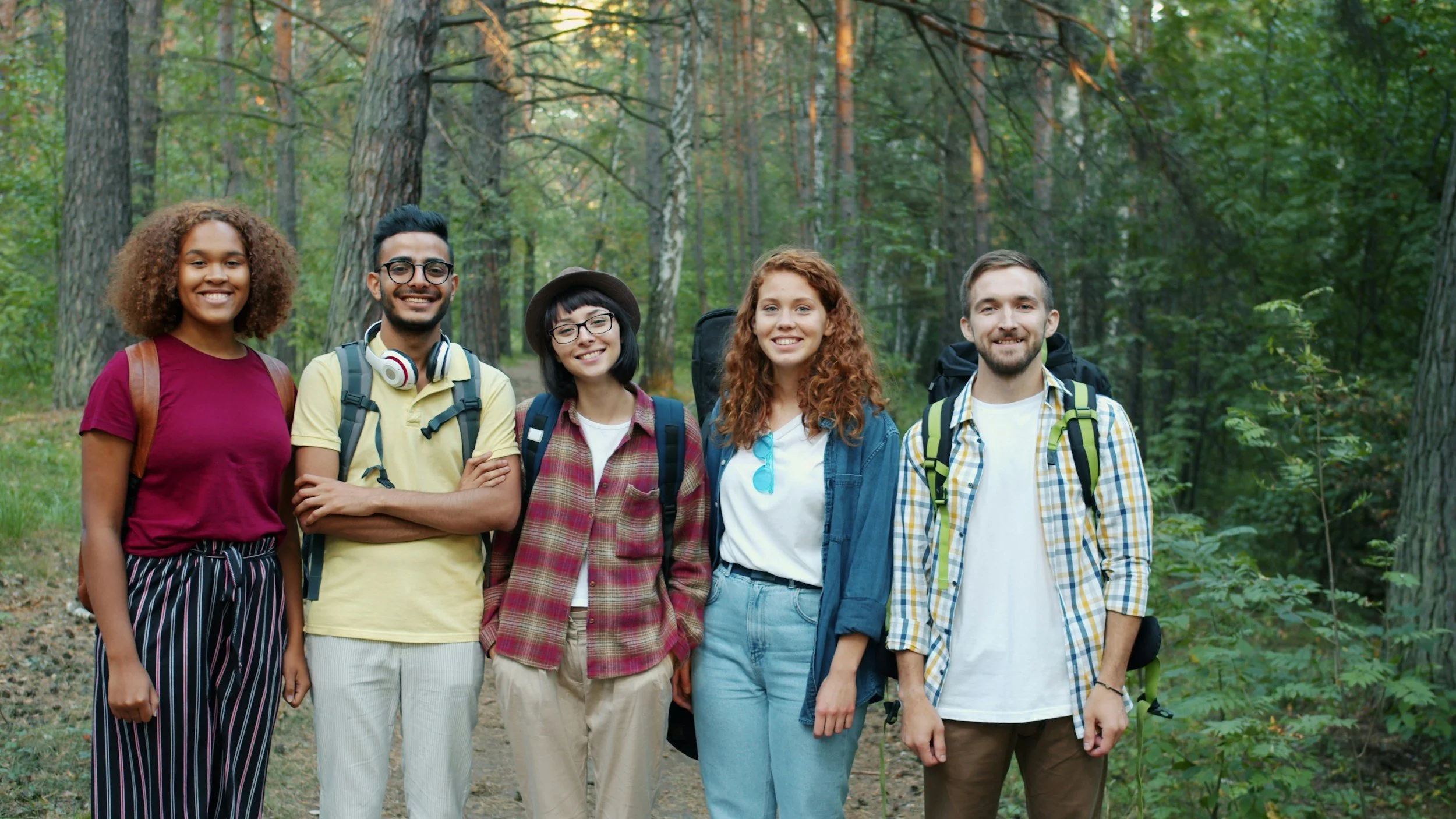 Five young adults standing together in a forest, smiling at the camera, with trees and greenery around them.