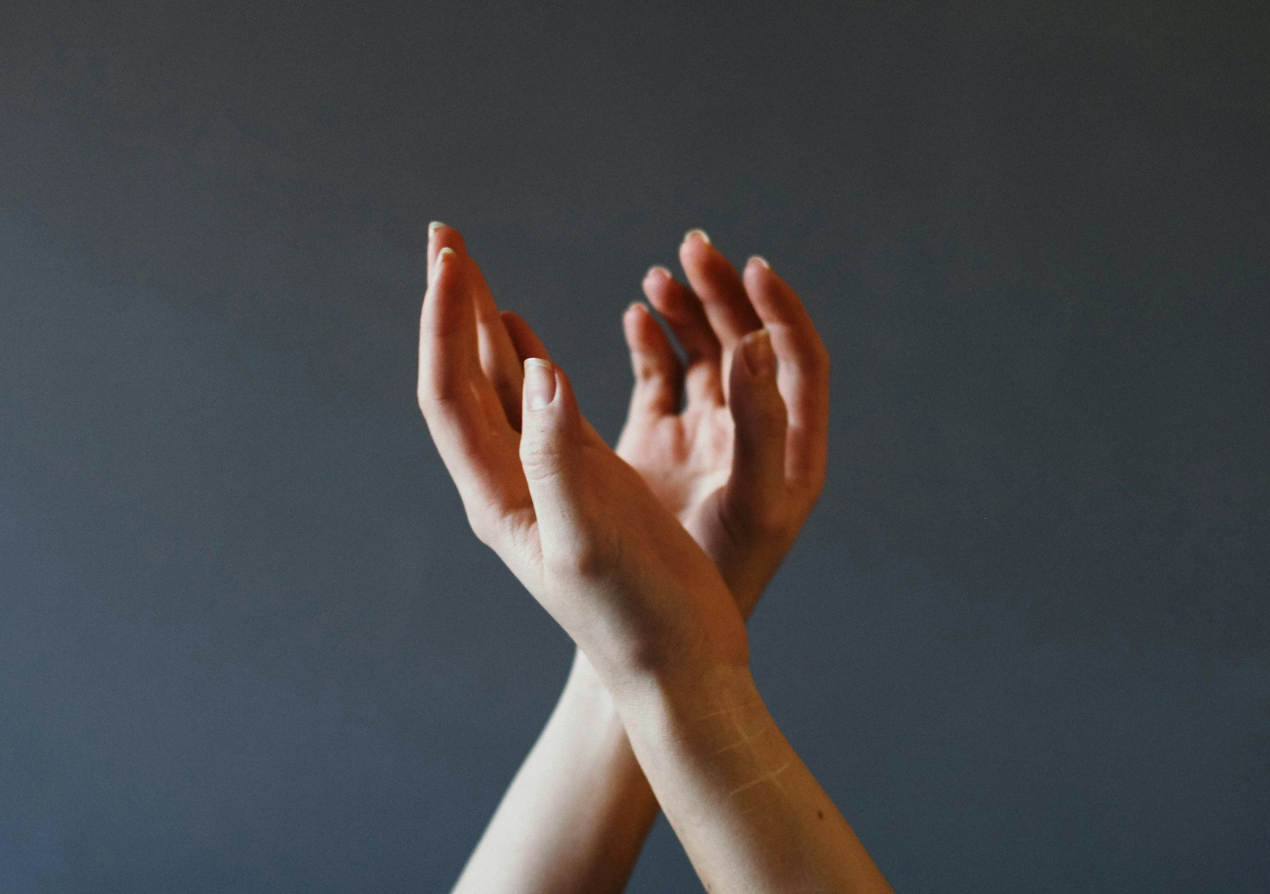 Two hands reaching towards the ceiling against a dark background, with one hand's fingers touching the other's palm.