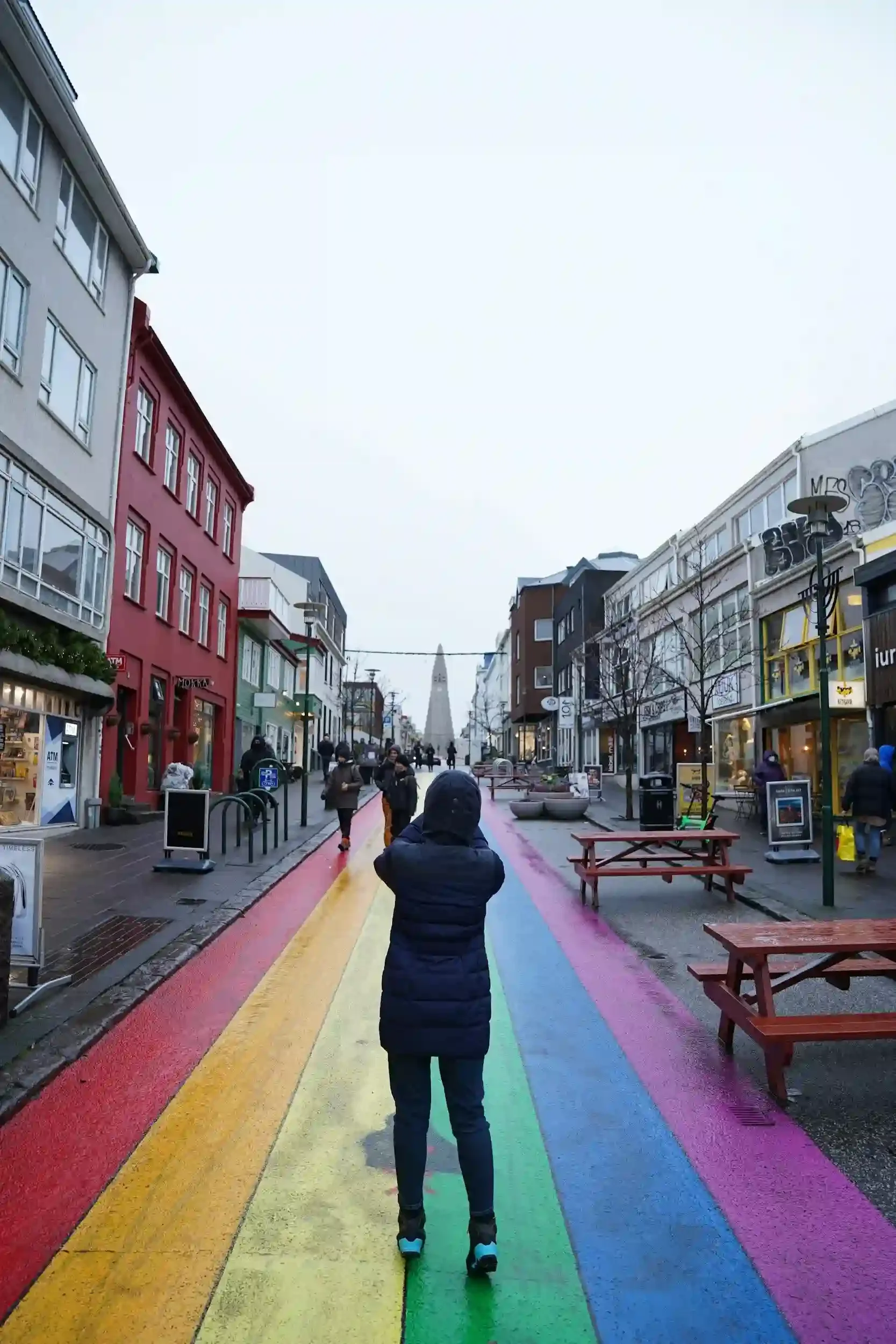 A person stands on the rainbow-painted street in Reykjavík, Iceland, looking toward the Hallgrímskirkja church under a cloudy sky.