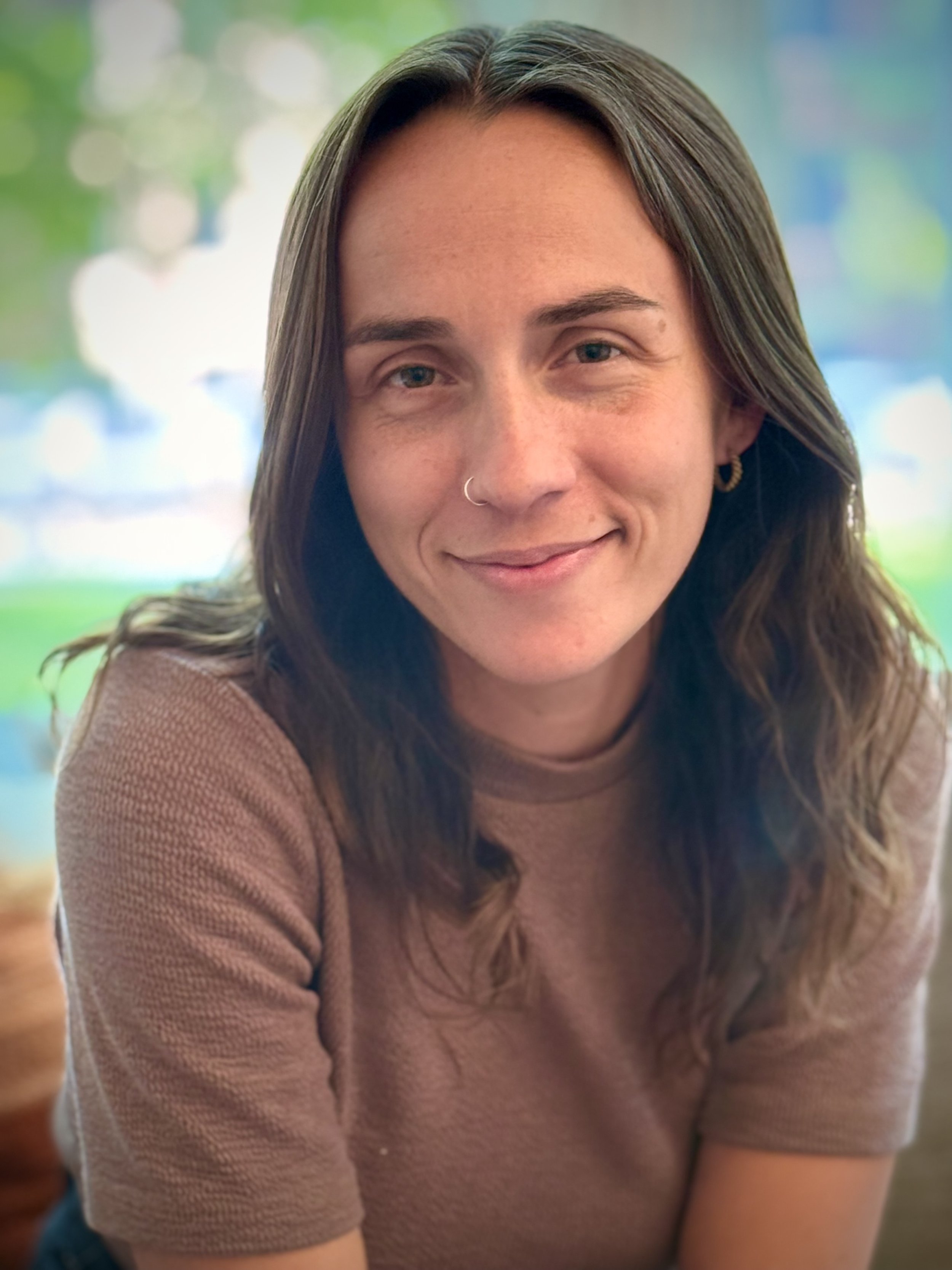 A young woman with long brown hair, wearing a nose ring and a short-sleeved beige shirt, smiling outdoors with a blurred background of trees and greenery.