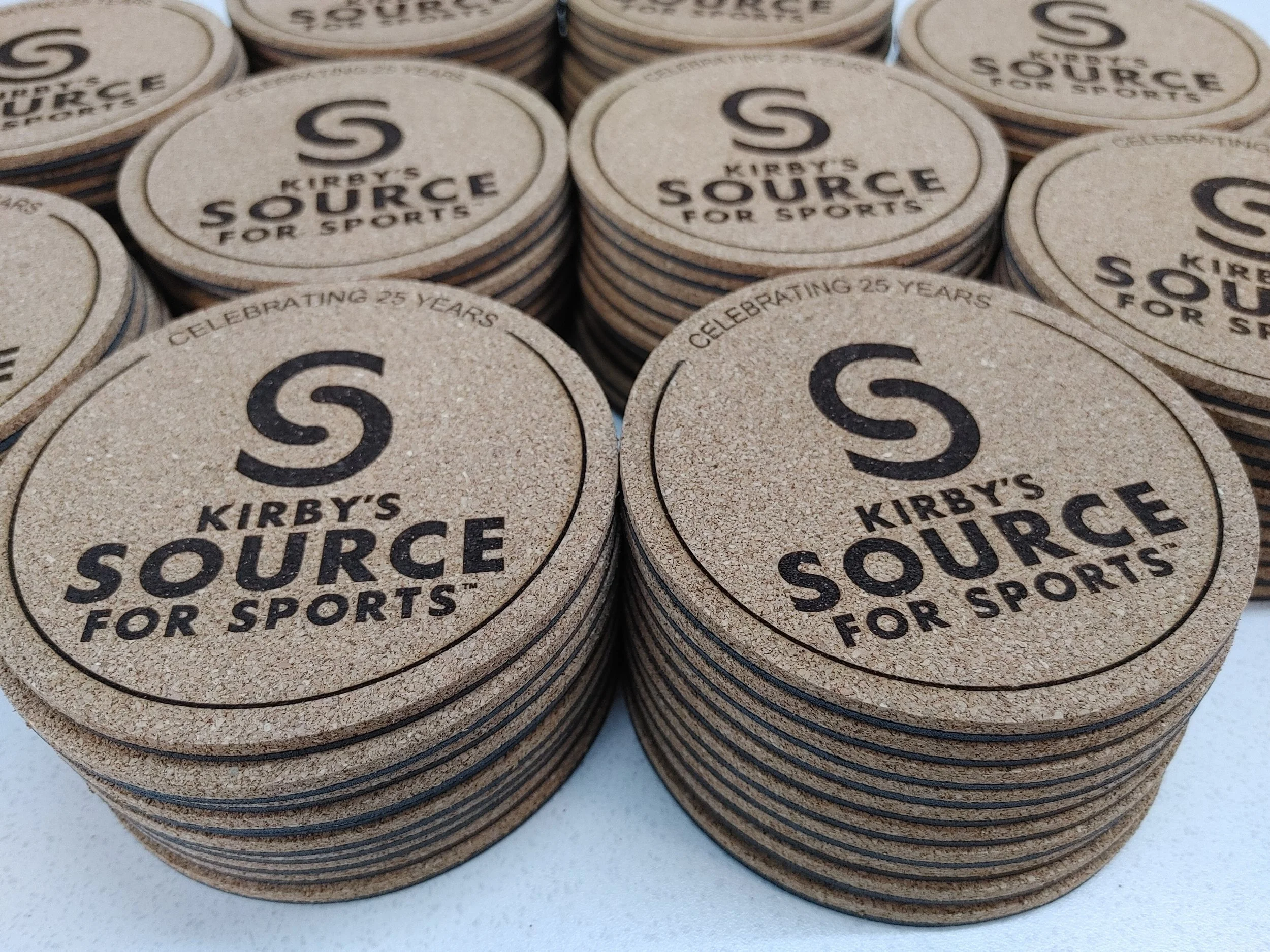 Stacked circular cork coasters with laser engraved black text reading 'KIRBY'S SOURCE FOR SPORTS' and 'CELEBRATING 25 YEARS' at the top edge, arranged in rows on a white surface.