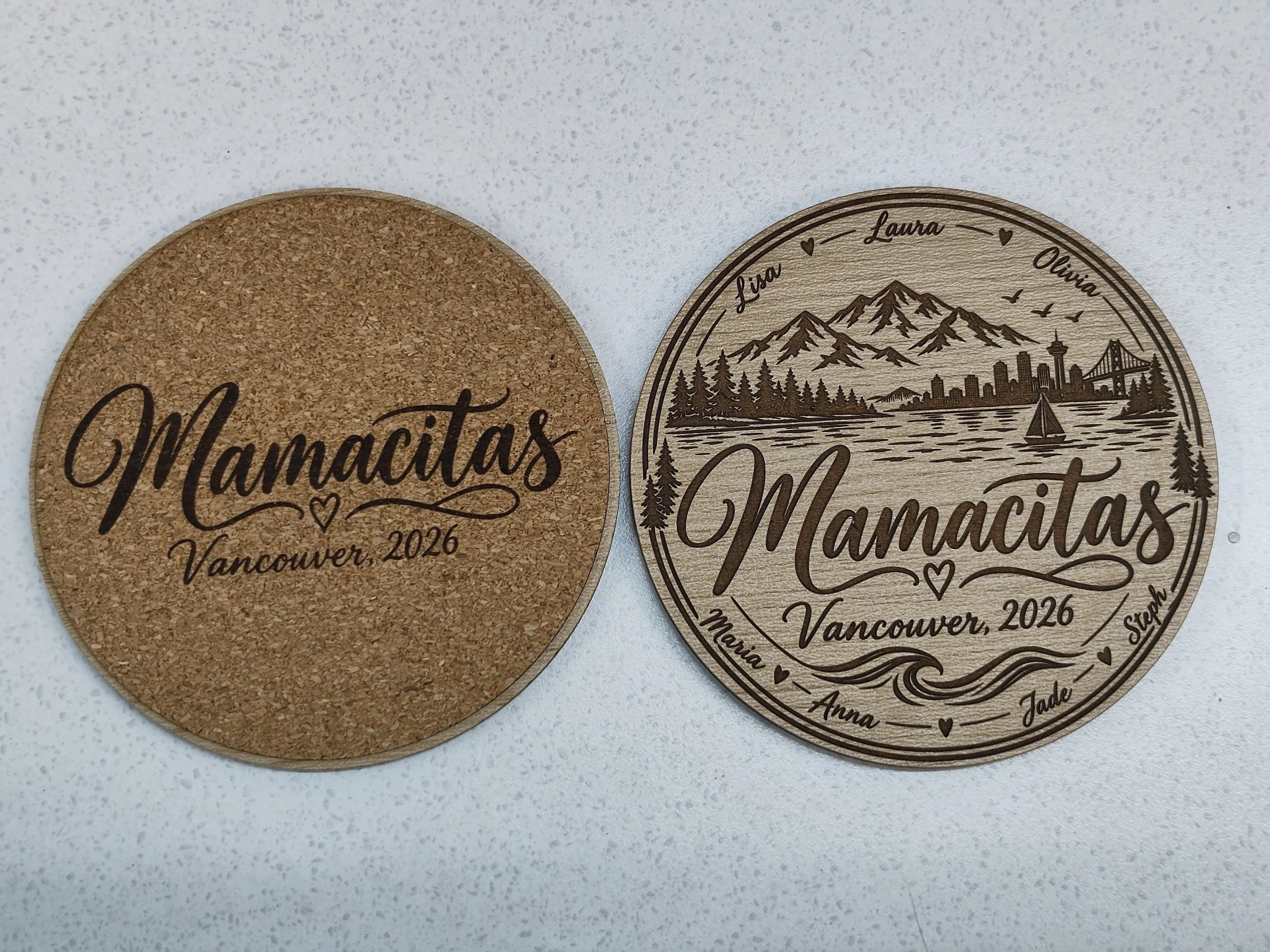 Two round wooden coasters, one with a cork surface and the other engraved with a mountain landscape and cityscape, with names 'Lisa,' 'Laura,' 'Olivia,' 'Jade,' and 'Stephen,' along with 'Vancouver 2026' and decorative hearts.