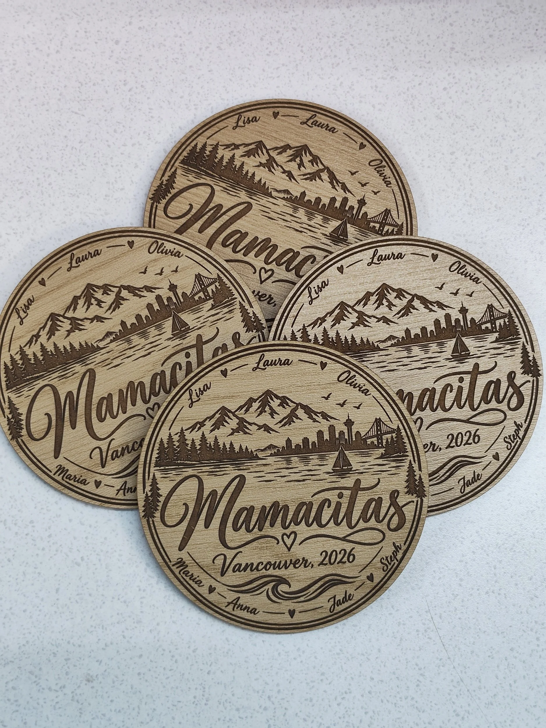 Wooden coasters with engraved images of mountains, a city skyline, a bridge, trees, a sailboat, and birds, personalized with the names Laura, Olivia, Lucas, and others, and labeled 'Vancouver, 2026.'
