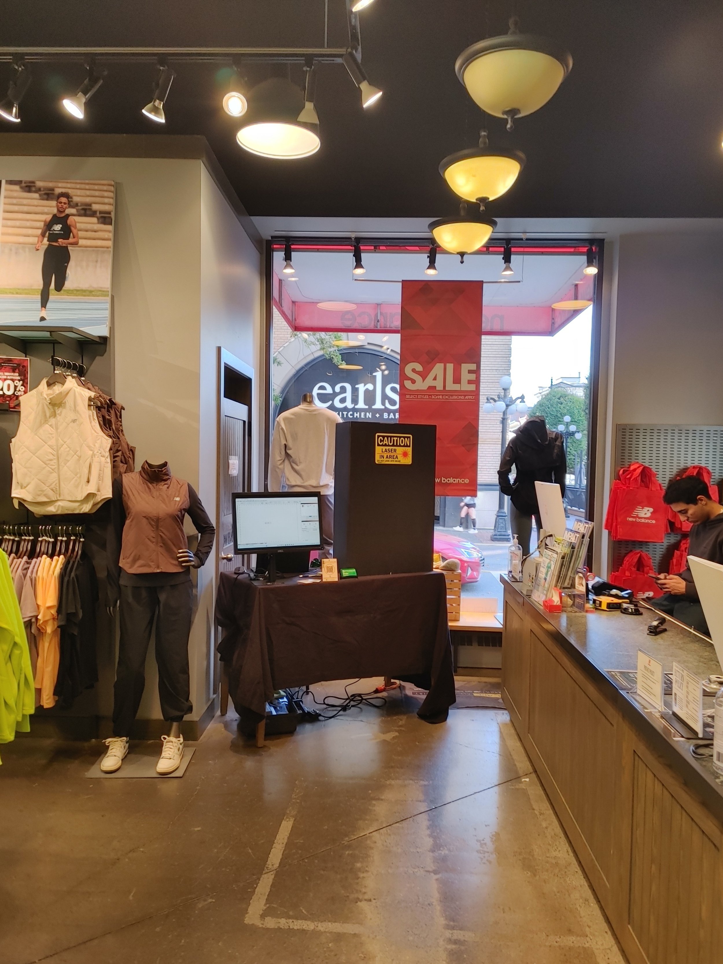 Inside of a retail sportswear store featuring mannequins dressed in athletic clothing, a computer workstation, a prominent sale sign, and a cashier counter. The background shows an outside view through the store's glass windows.